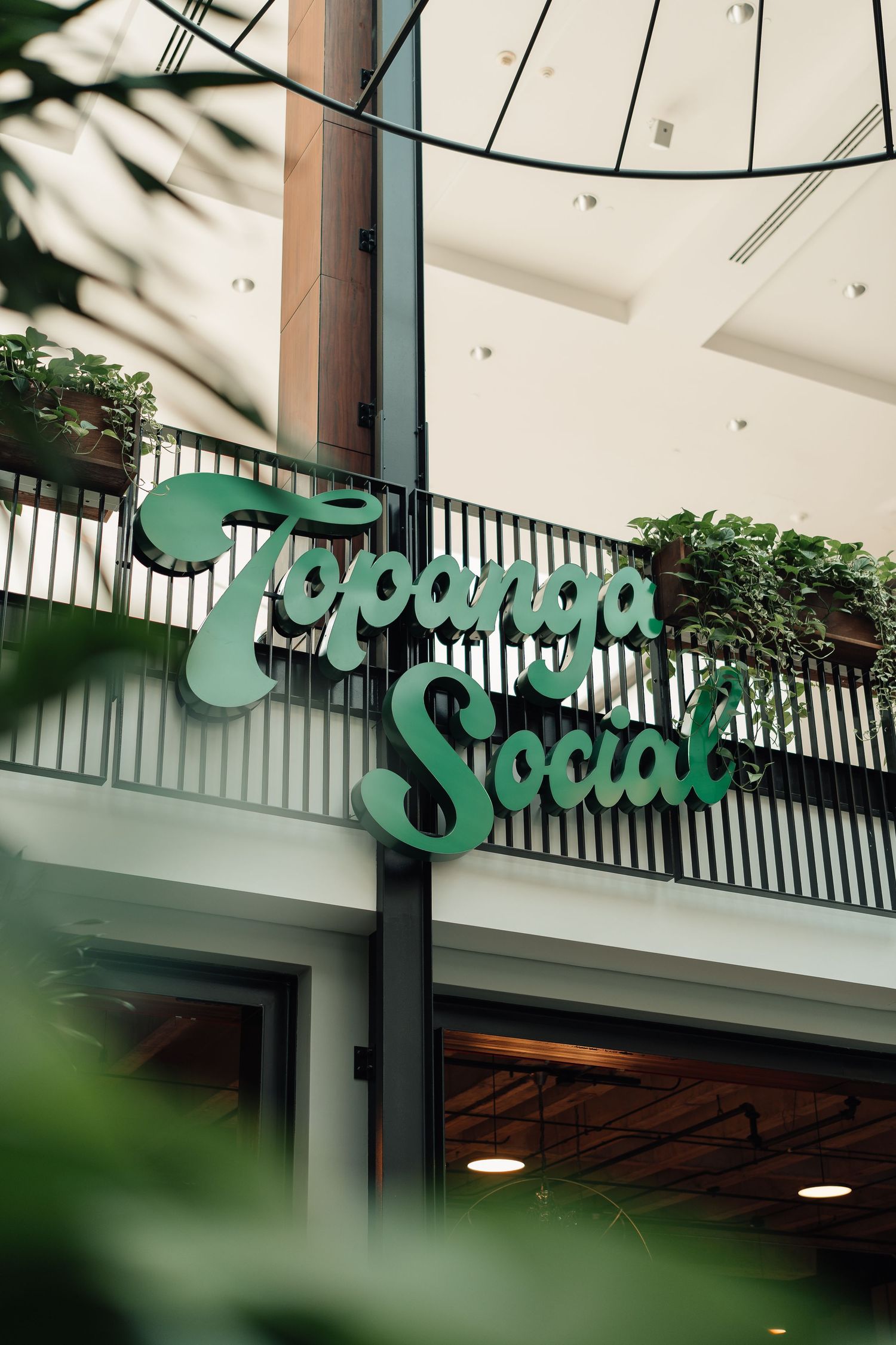 Green retro script Topanga Social sign mounted on a pillar inside a modern shopping mall.