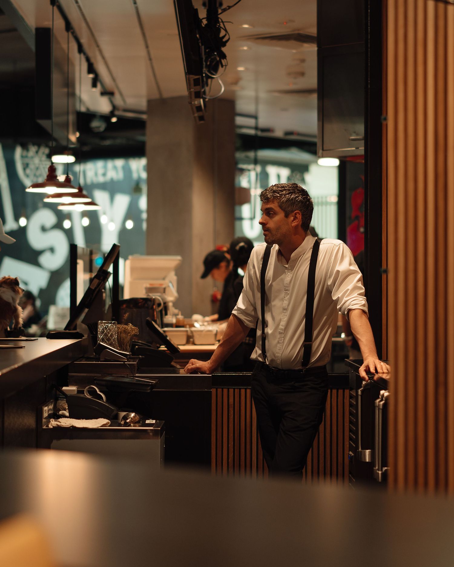 Man in white shirt and suspenders working at a restaurant counter under warm pendant lights.