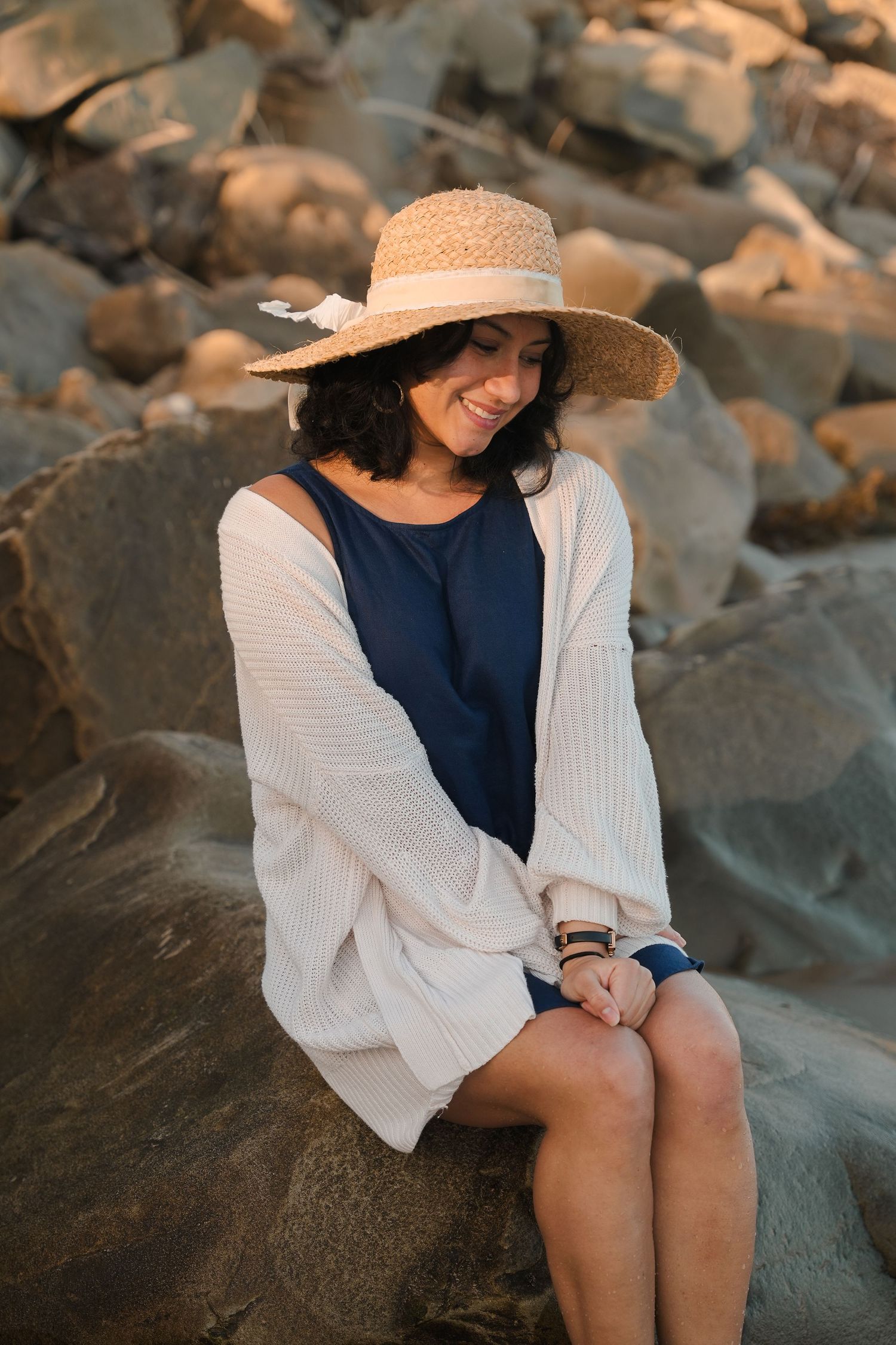 Smiling woman in straw hat and cream cardigan sitting on rocks at golden hour.