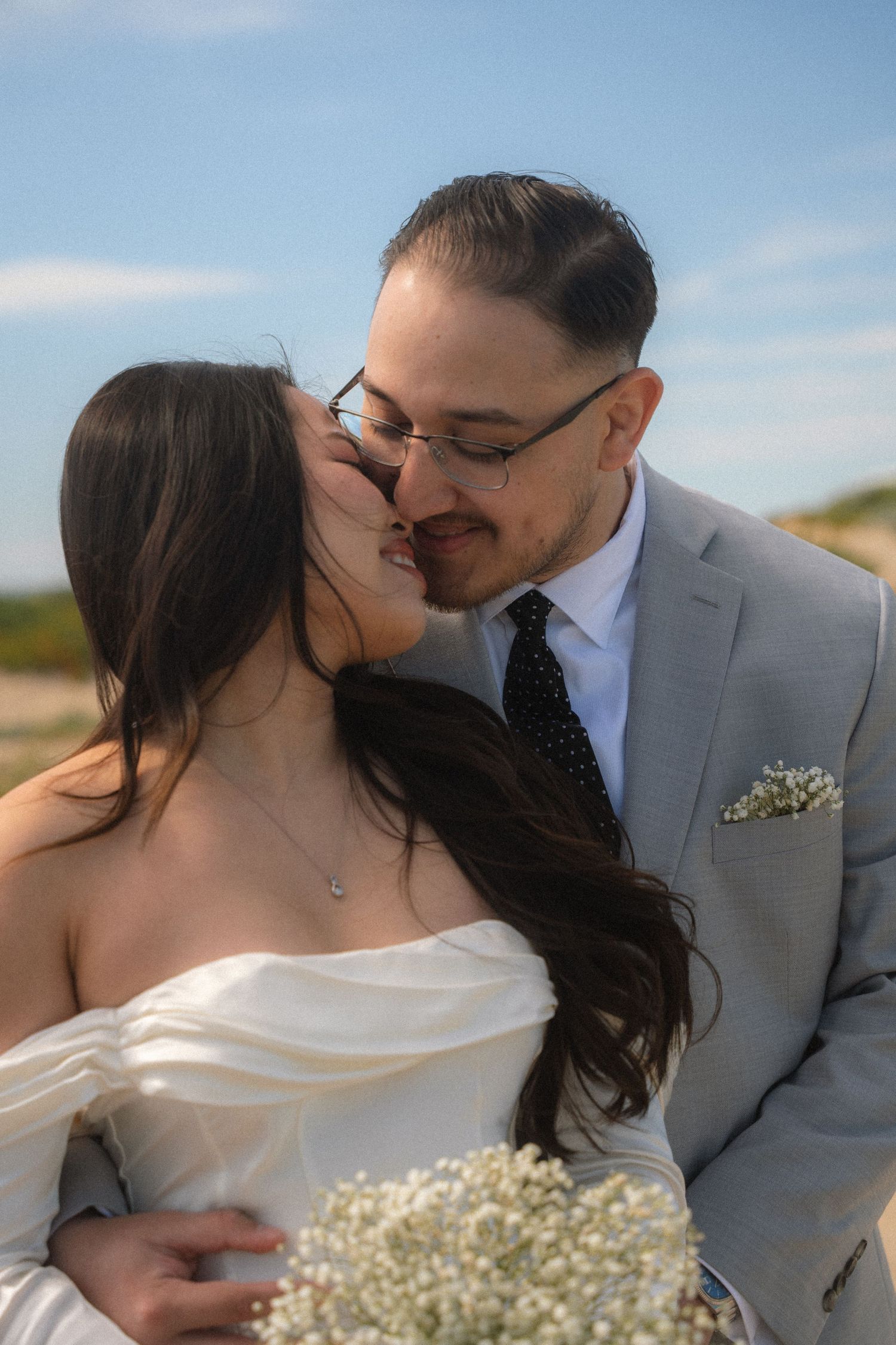 Groom in gray suit kisses bride in white off-shoulder dress outdoors on a sunny day.