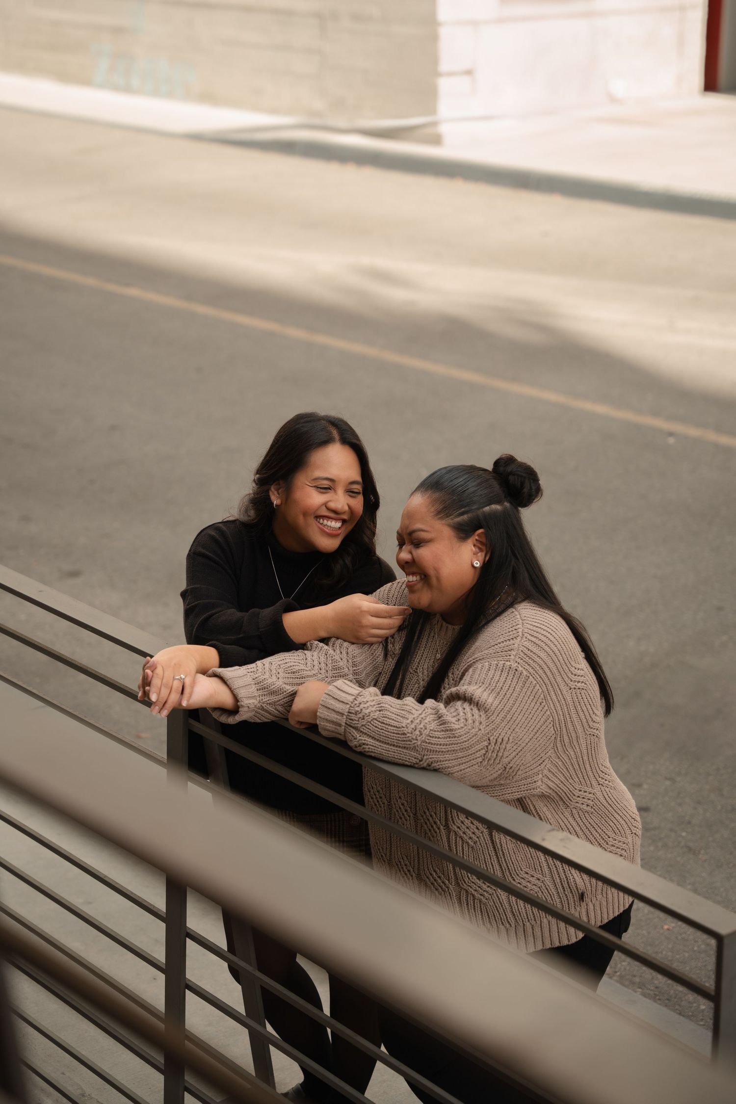 Two women laughing and leaning on a railing outdoors on a city street.