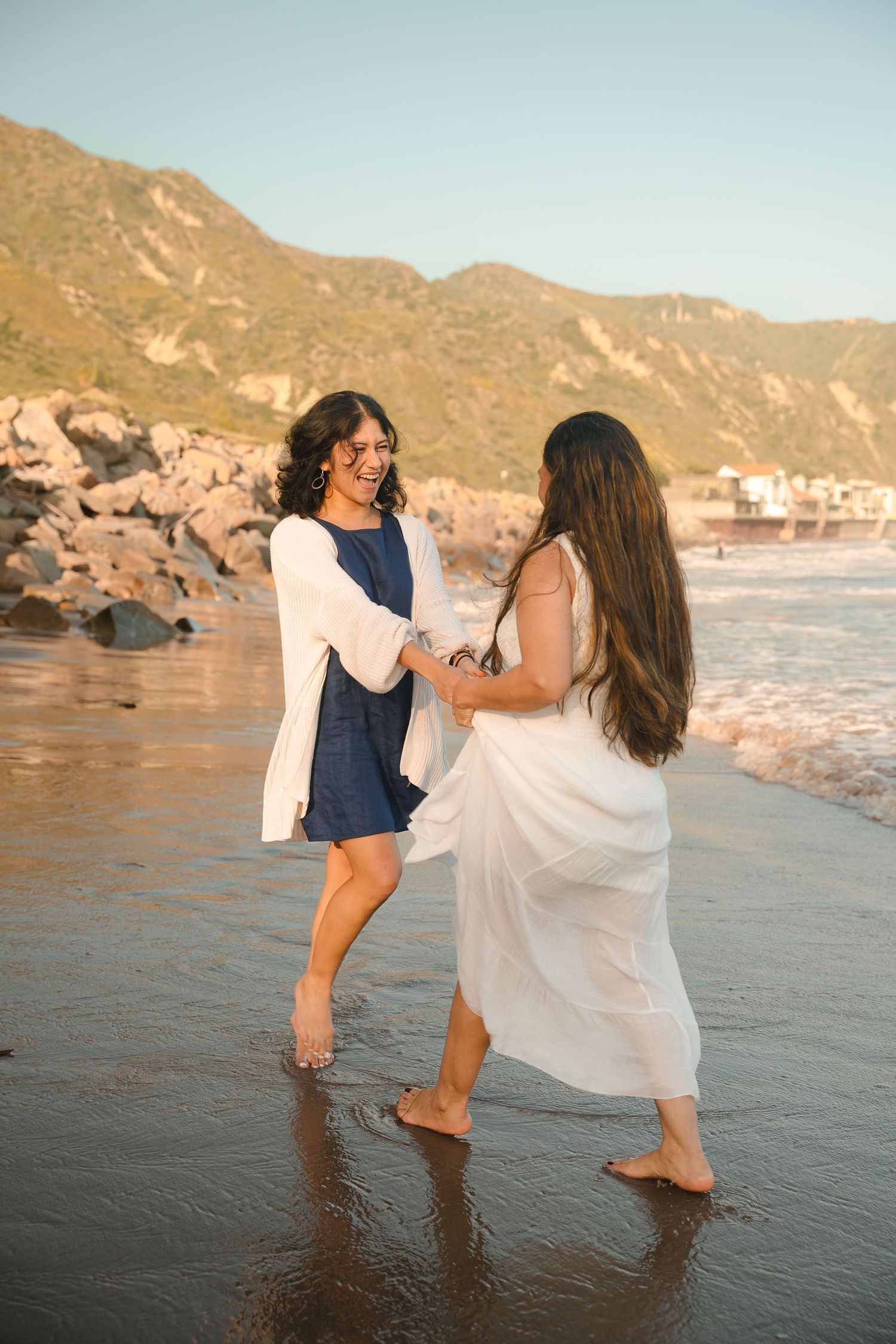 Two women laughing and holding hands while walking barefoot on a sunny beach.