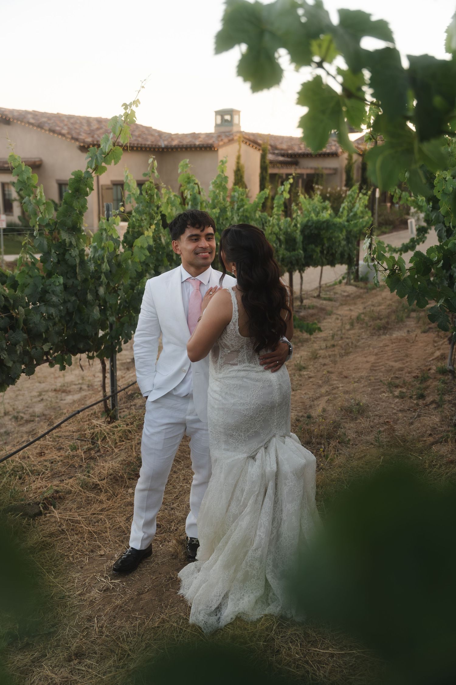 Smiling groom in white suit with bride in lace dress among vineyard rows at sunset.