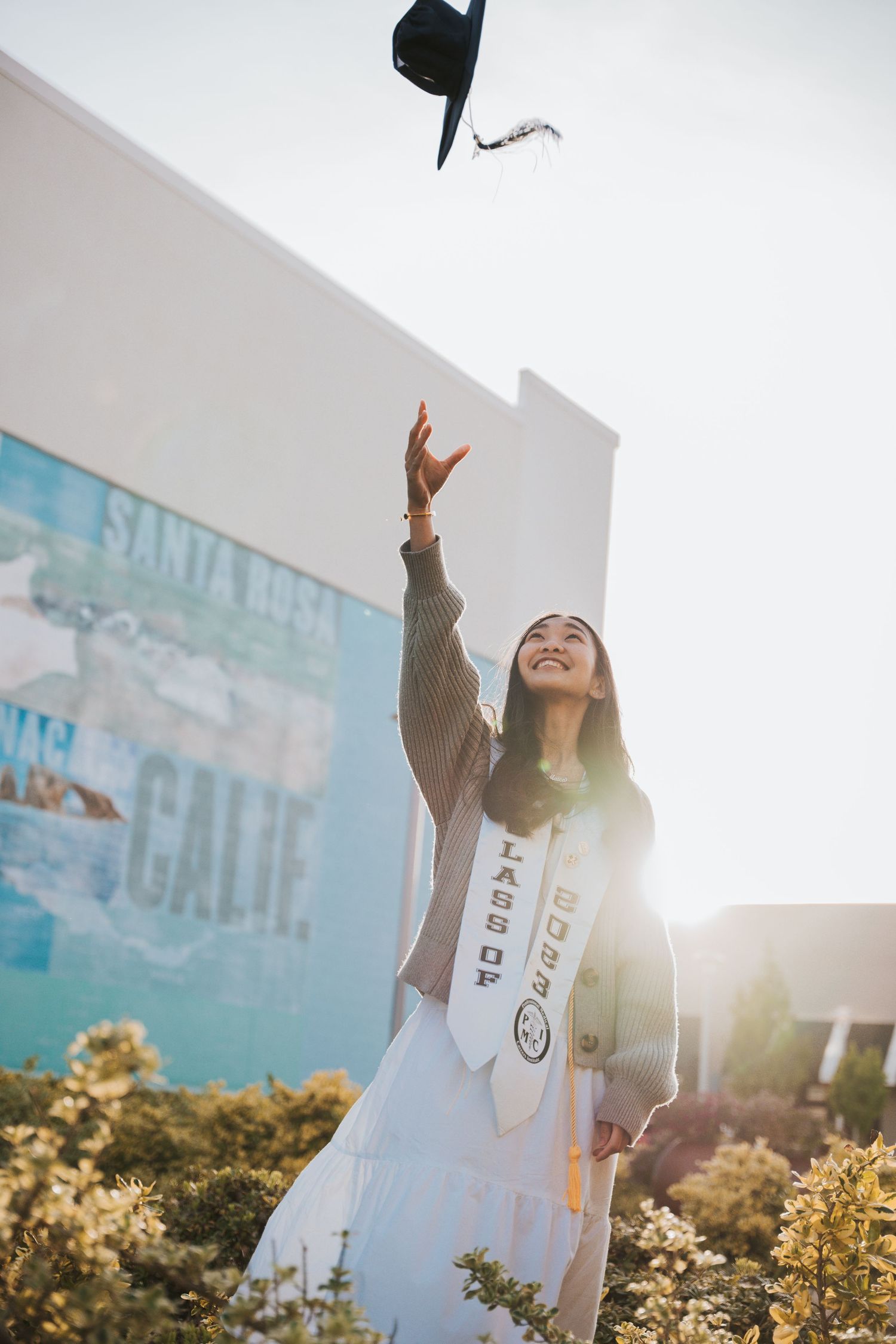 Graduate in Class of 2023 stole tossing cap in air near Santa Rosa California mural in sunlight.