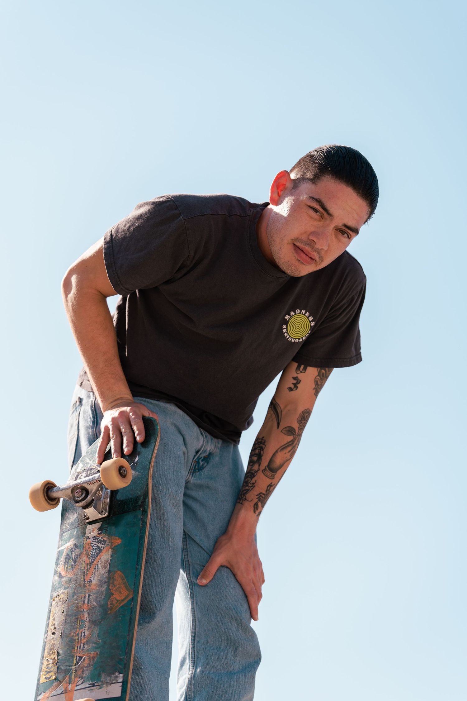 Tattooed young man leaning forward holding a skateboard against a clear blue sky.