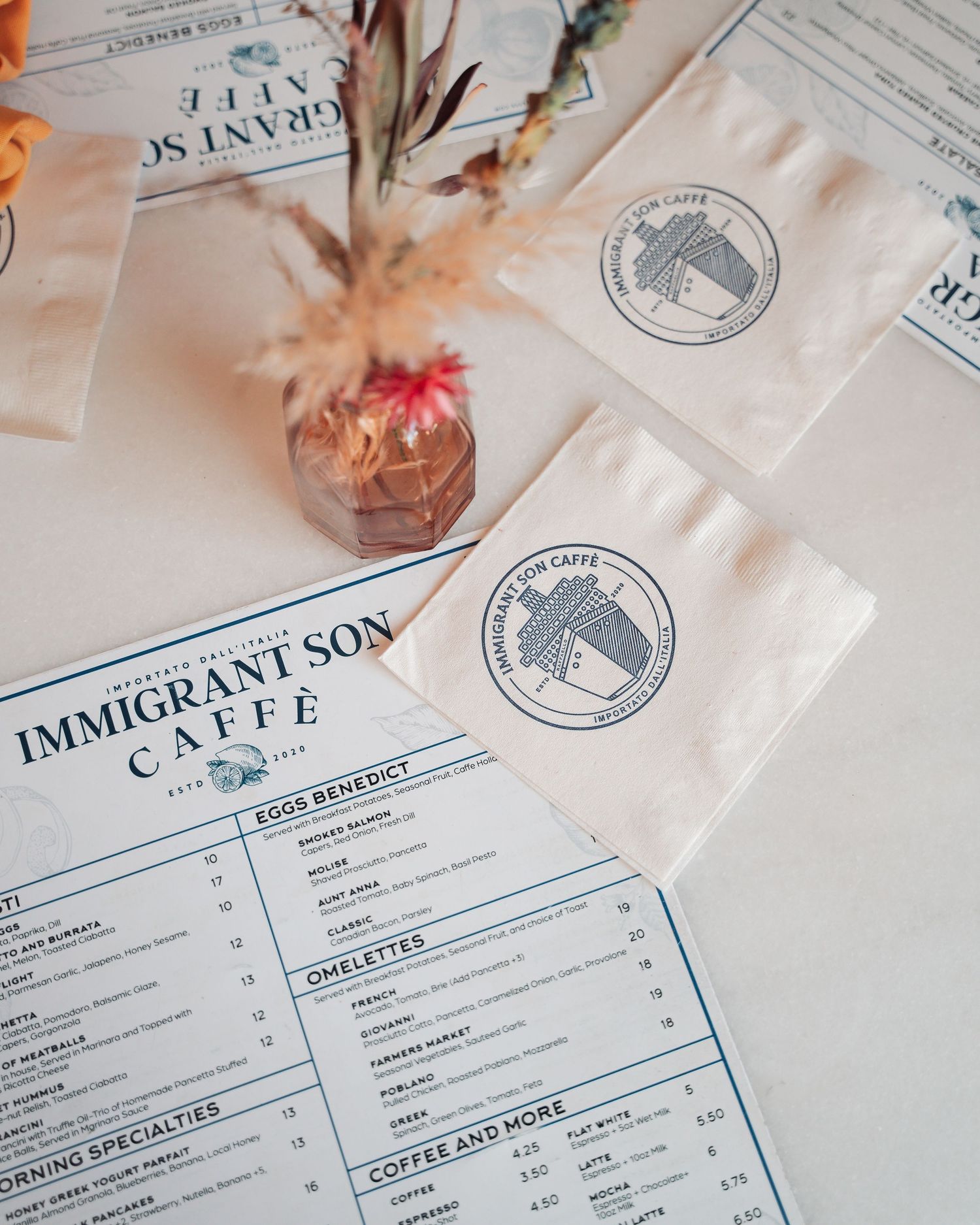 Immigrant Son Caffe menu with branded napkins and dried floral centerpiece on white tablecloth.