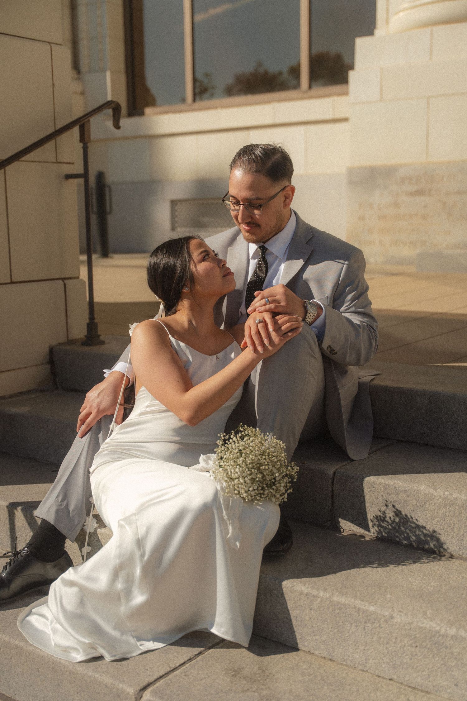 Bride and groom sitting on stone steps, holding hands with small white bouquet nearby.
