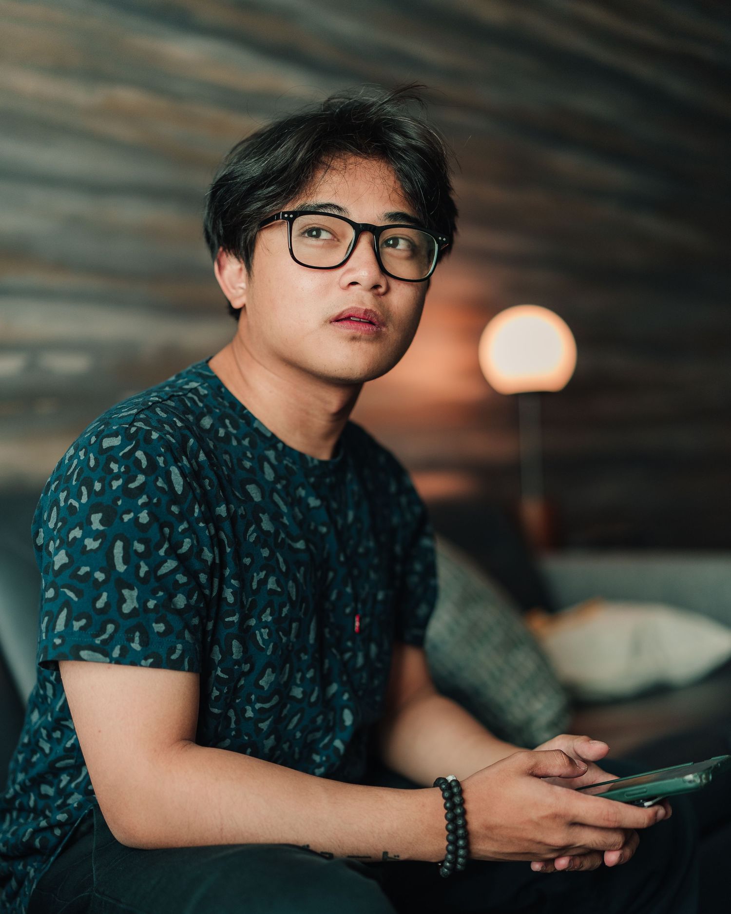 Young man with glasses holding a phone, sitting indoors with warm ambient lighting behind him.