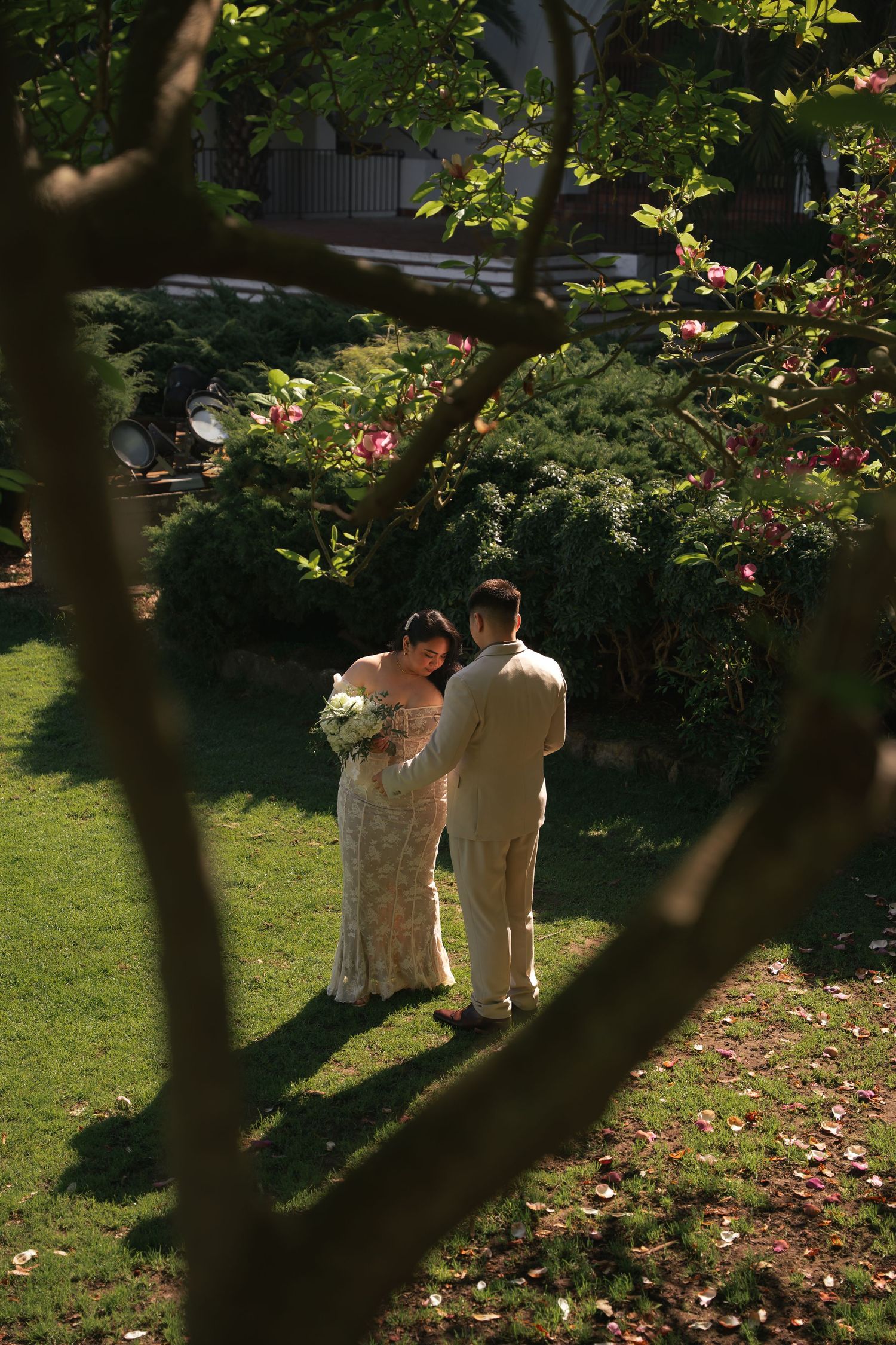 Bride and groom sharing intimate moment in lush garden, viewed through blooming tree branches.