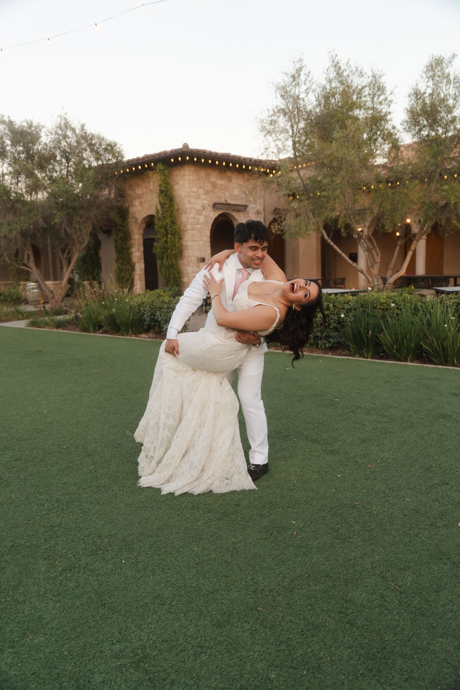 Groom dipping bride in white lace gown on green lawn outside rustic wedding venue.