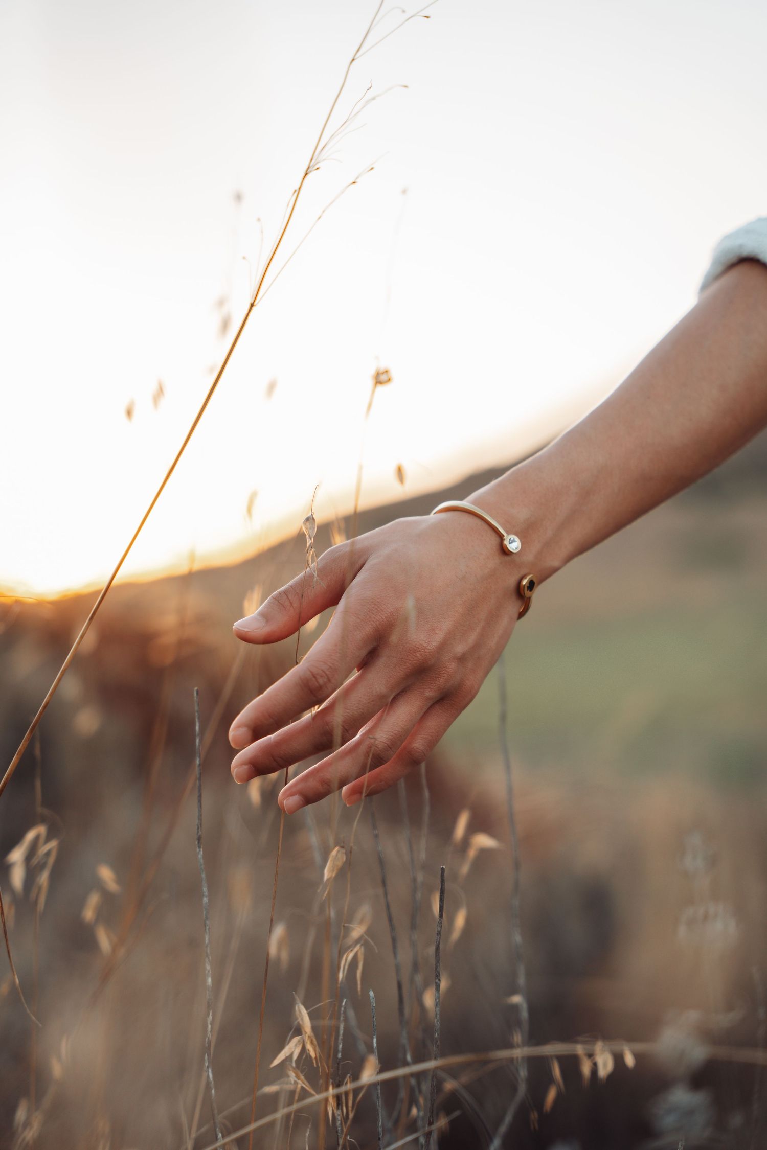 Close-up of a womans hand with a gold bracelet gently touching tall grass at sunset.