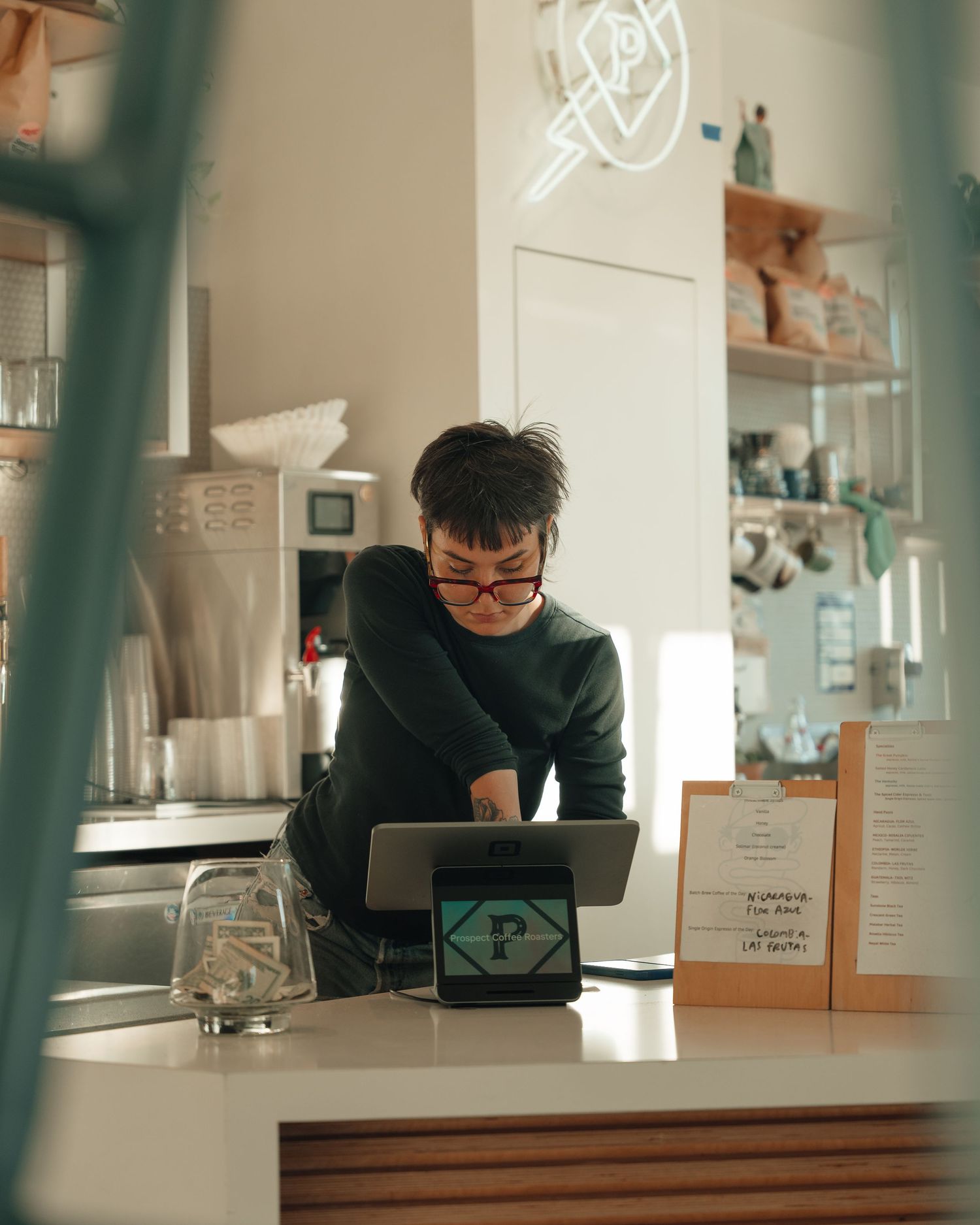 Barista in green sweater operating tablet POS system at modern white coffee shop counter.