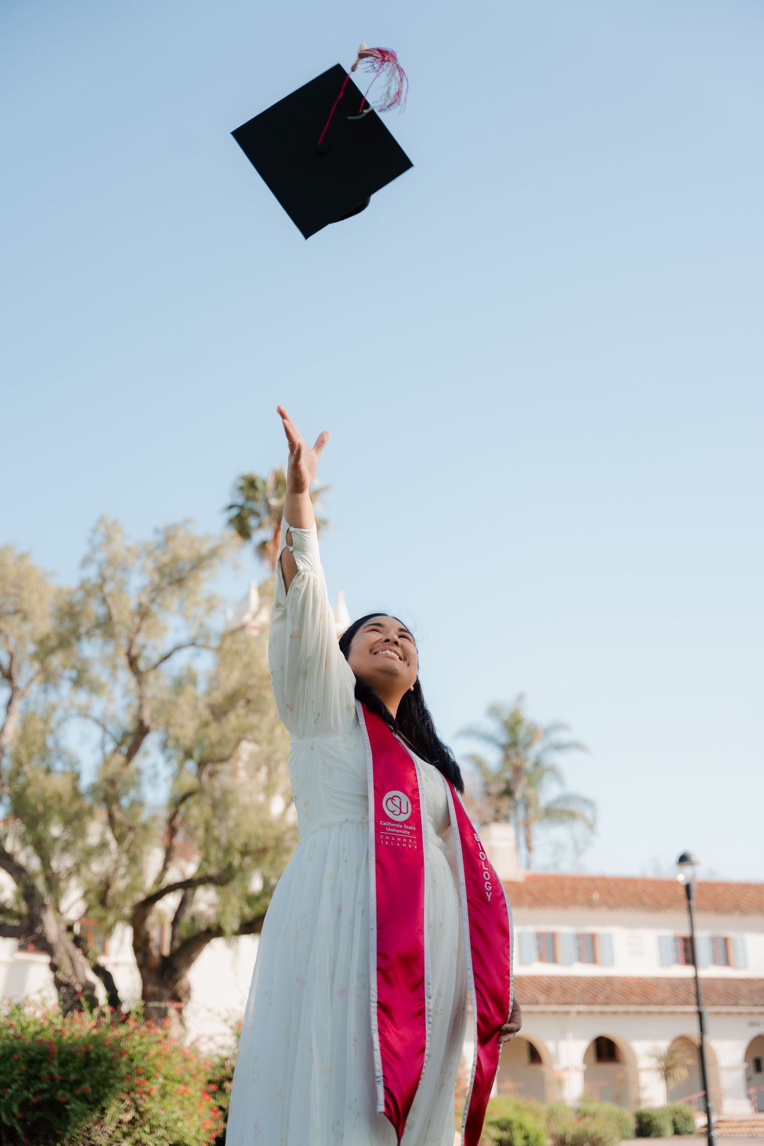 Smiling graduate in white gown tosses cap into blue sky at outdoor graduation ceremony.