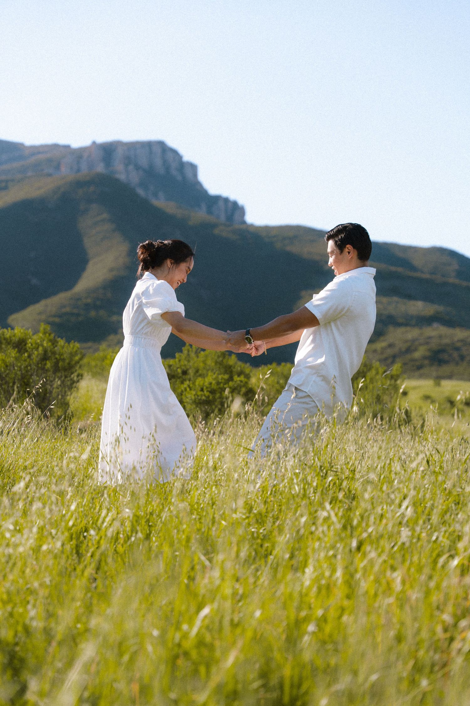 Joyful couple in white outfits holding hands in lush green meadow with mountain backdrop.