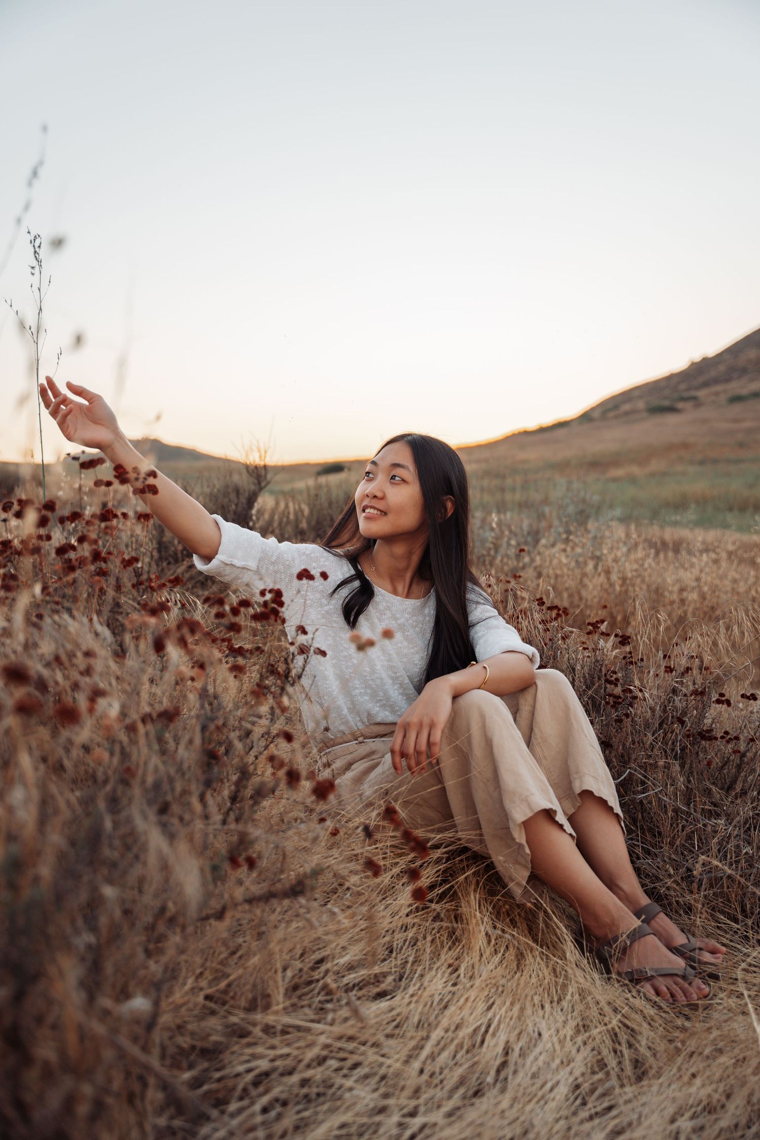 Young Asian woman sitting in dry grass field at sunset, reaching up to touch wildflowers.