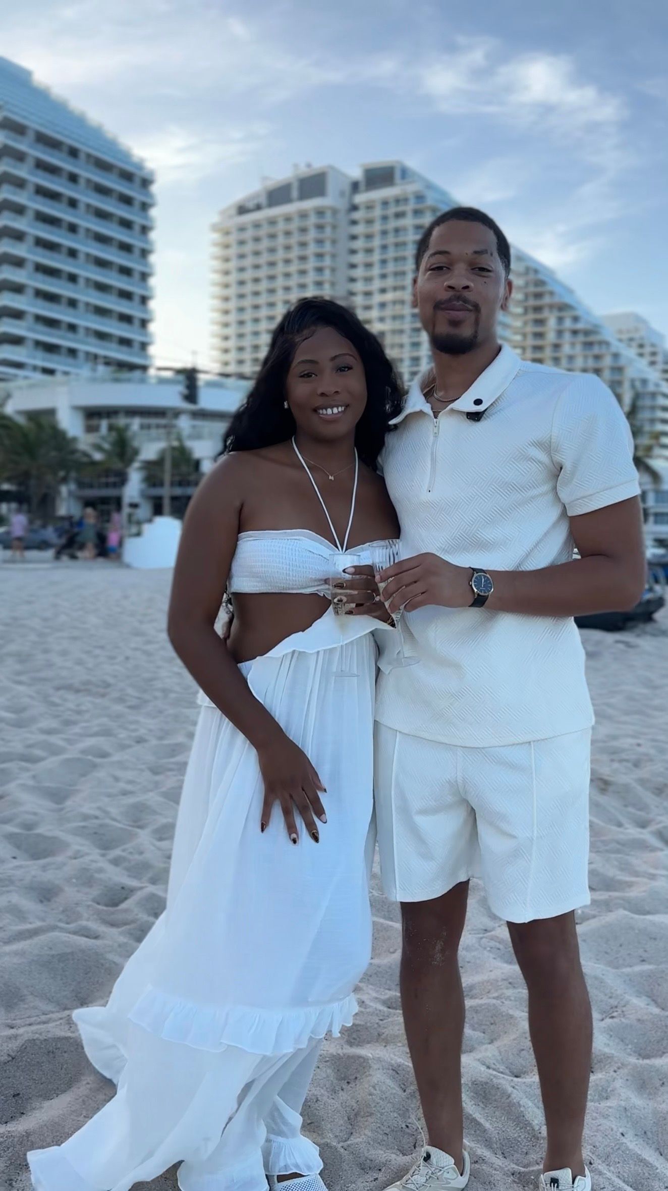 Couple in matching white outfits posing on a sandy beach with luxury hotels in the background at dusk.