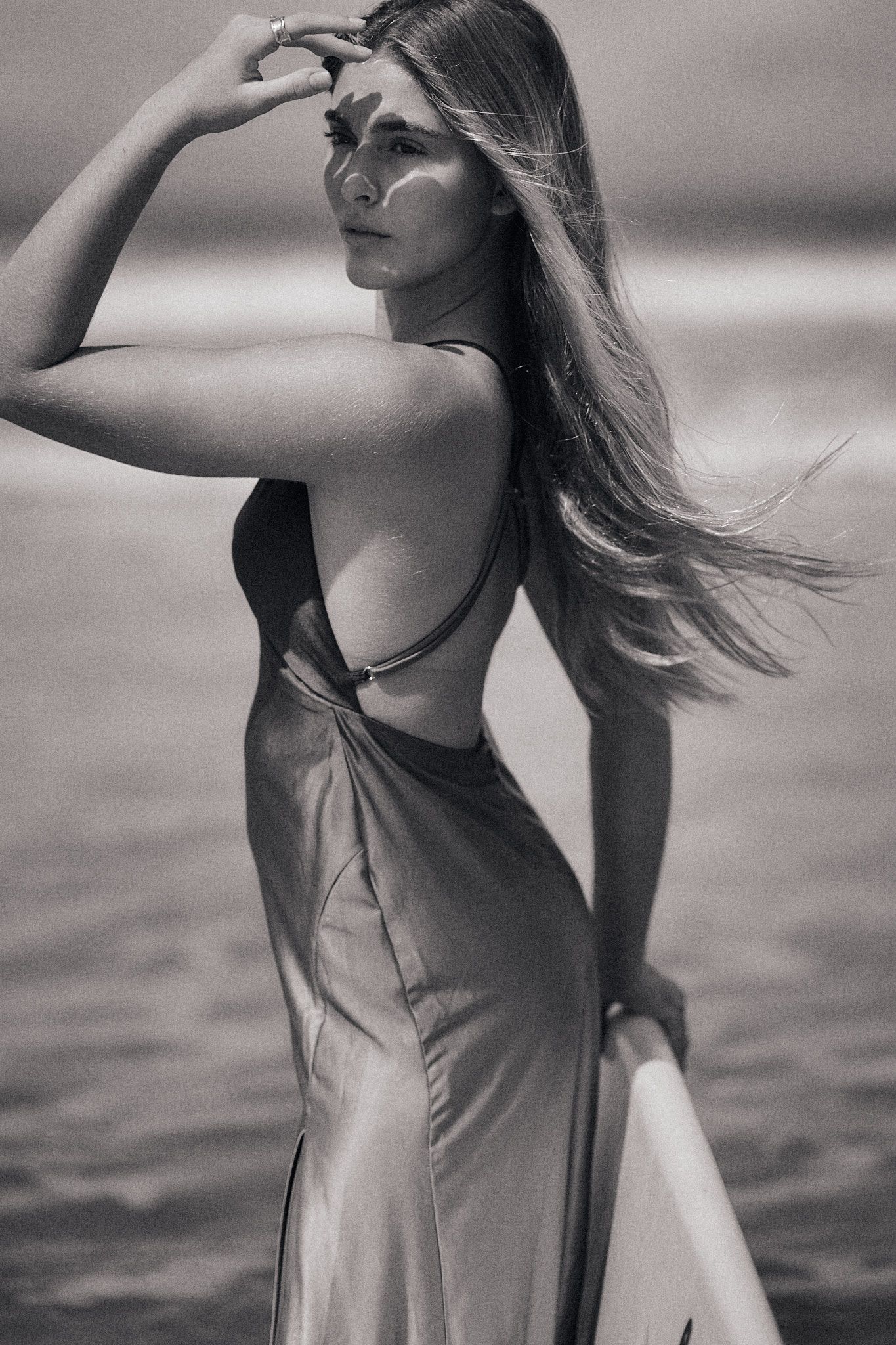 Black and white photo of a woman in a backless dress holding a surfboard on a beach with wind-blown hair.