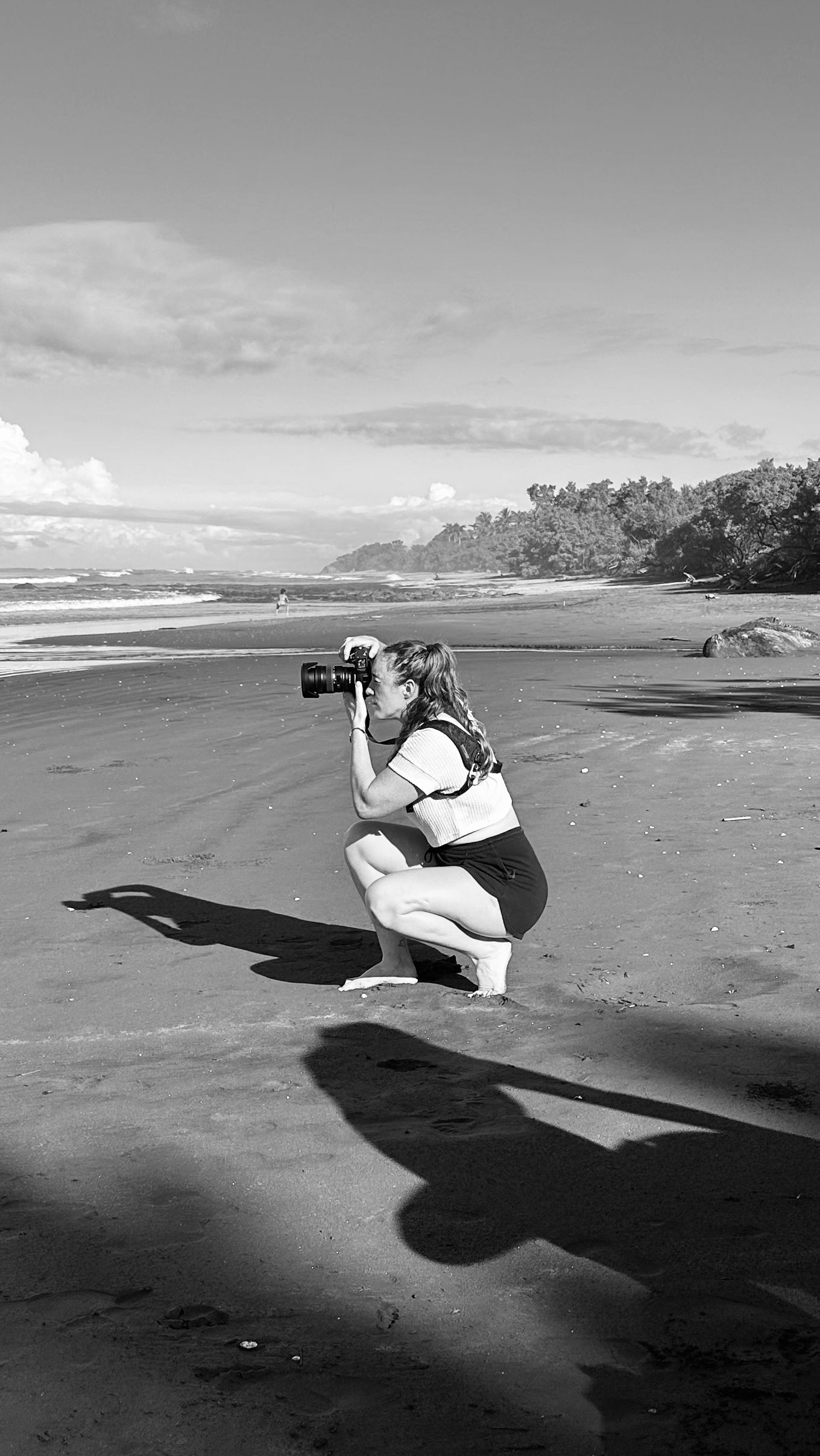 Black and white photo of a woman crouching on a beach, holding a camera up to take a photo, with ocean waves in the background.