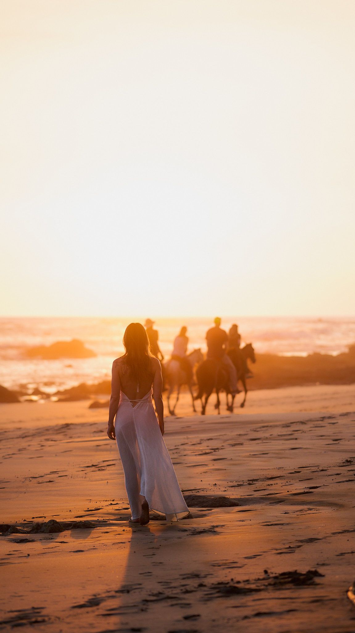 Person in white dress walking on beach at sunset while horse riders silhouetted against golden sky in background.