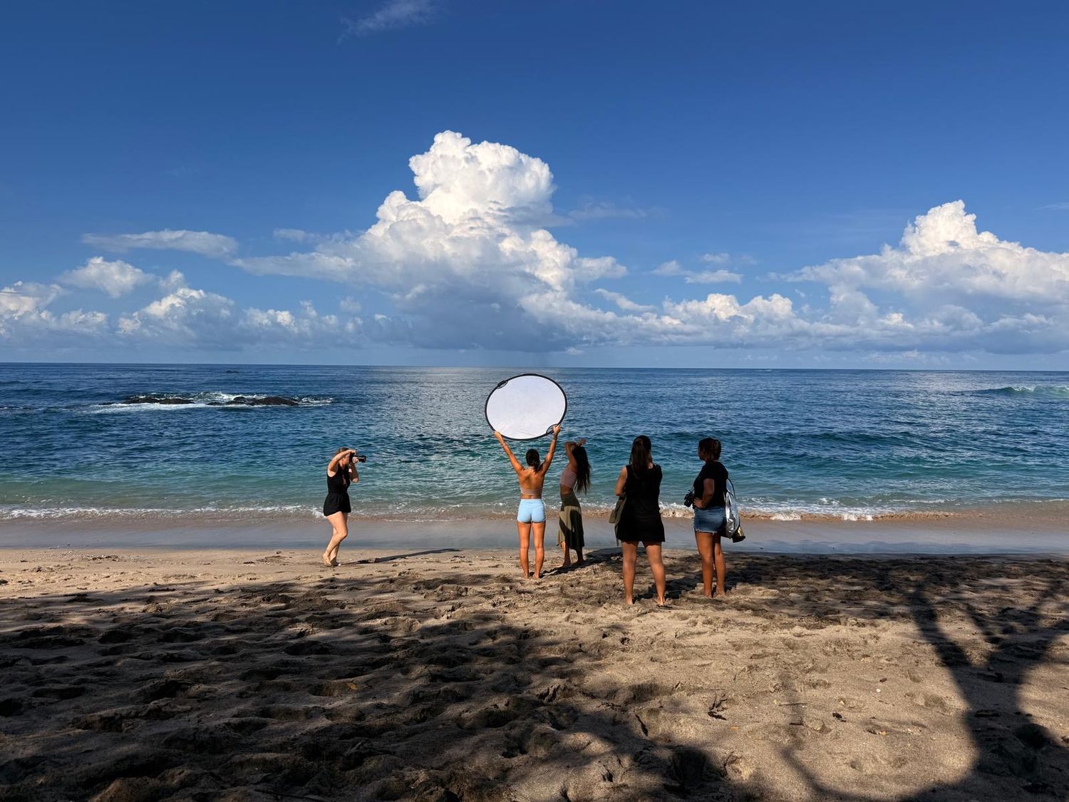 A photography crew with a reflector shoots models on a tropical sandy beach with turquoise water and dramatic clouds.