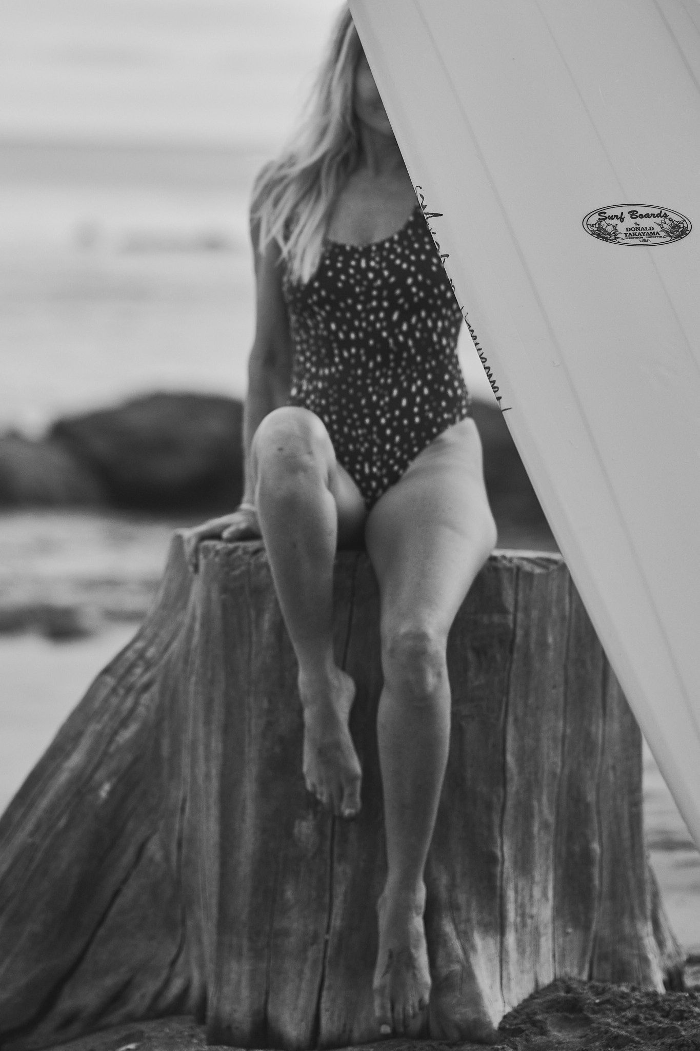 Black and white photo of someone in a polka dot swimsuit sitting on a wooden surface near a surfboard at the beach.