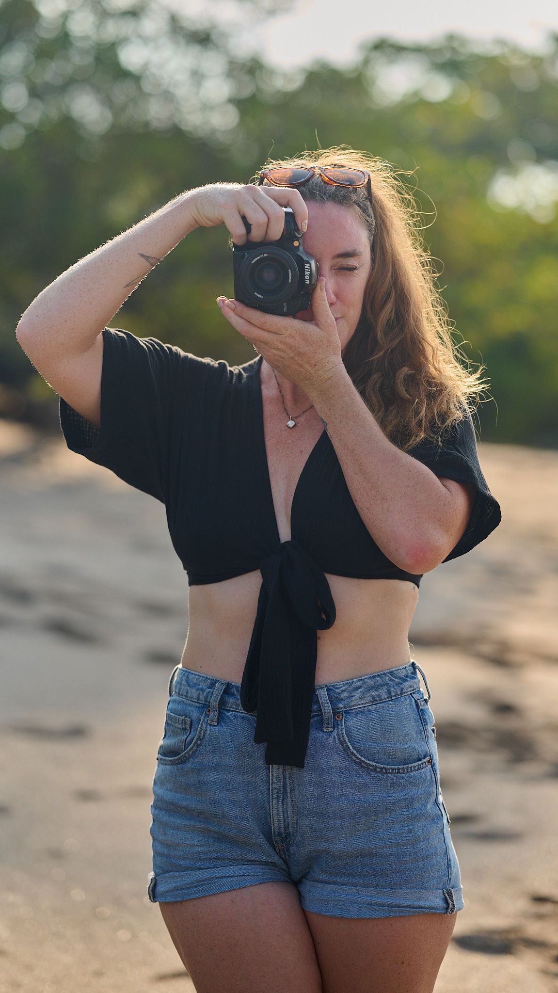 Woman in black crop top and denim shorts photographing outdoors with a DSLR camera at golden hour.