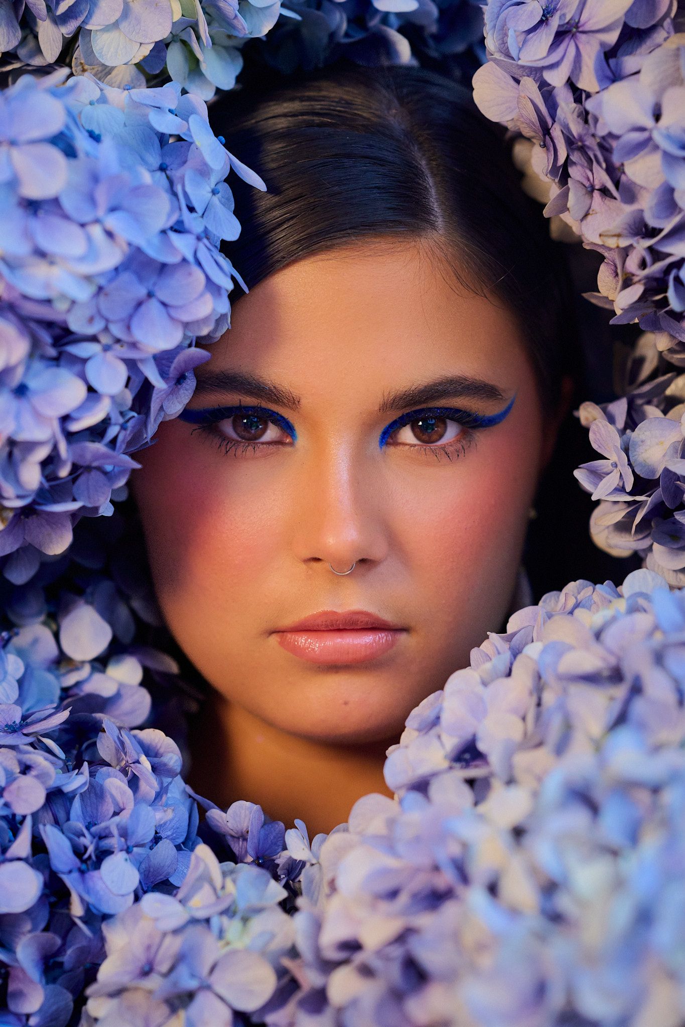 Beauty portrait surrounded by purple and blue hydrangea flowers with dramatic eye makeup and moody lighting.