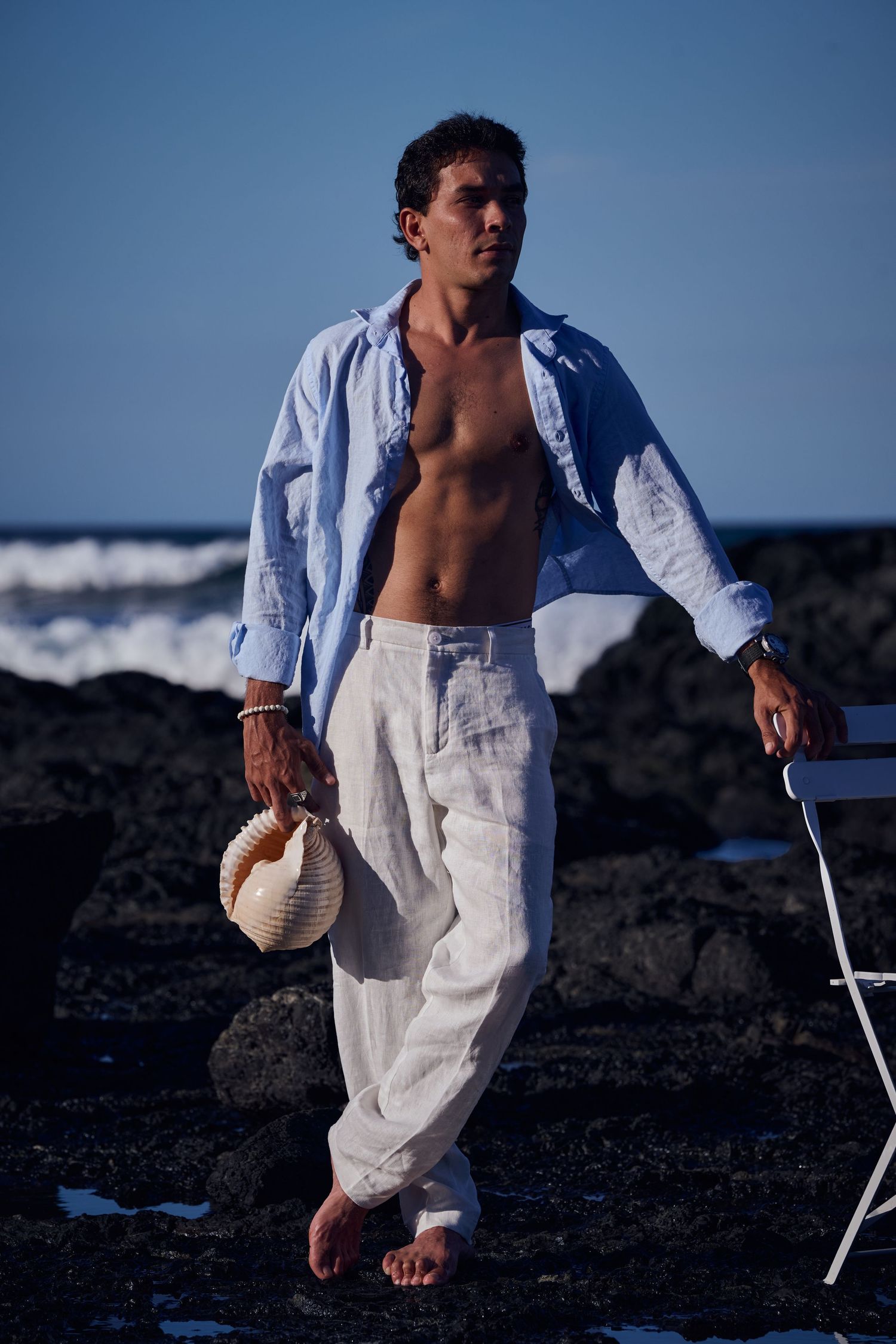 Shirtless man in white linen pants and open blue shirt walks barefoot on rocky beach holding a hat.