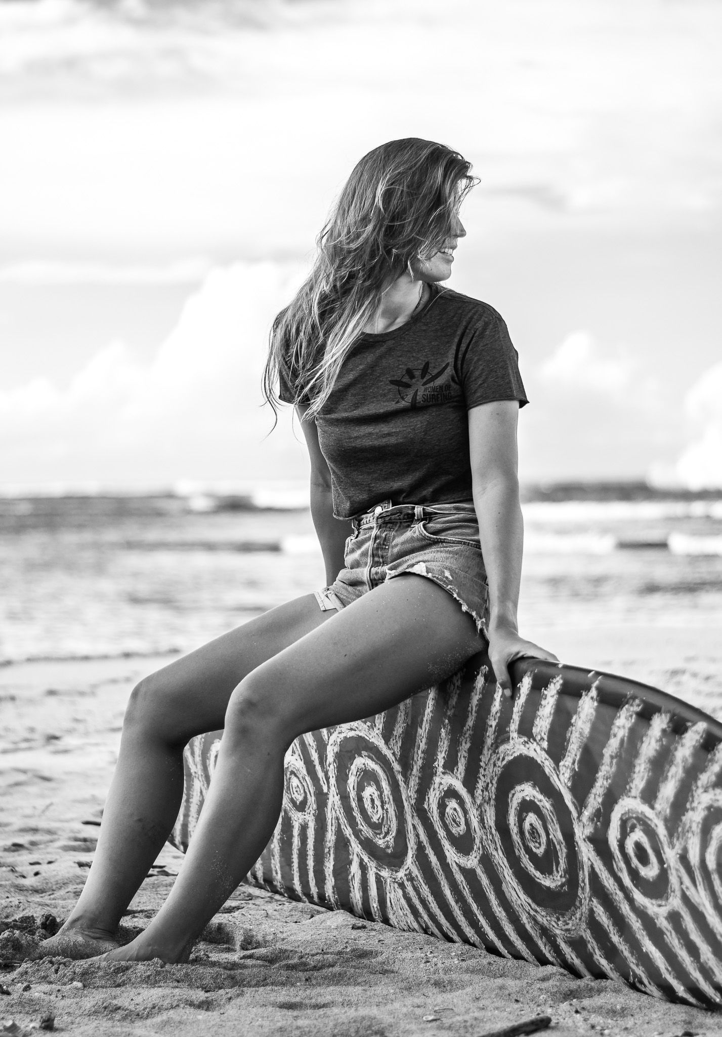 Black and white photograph showing a person sitting on a driftwood trunk at a beach with waves in the background.