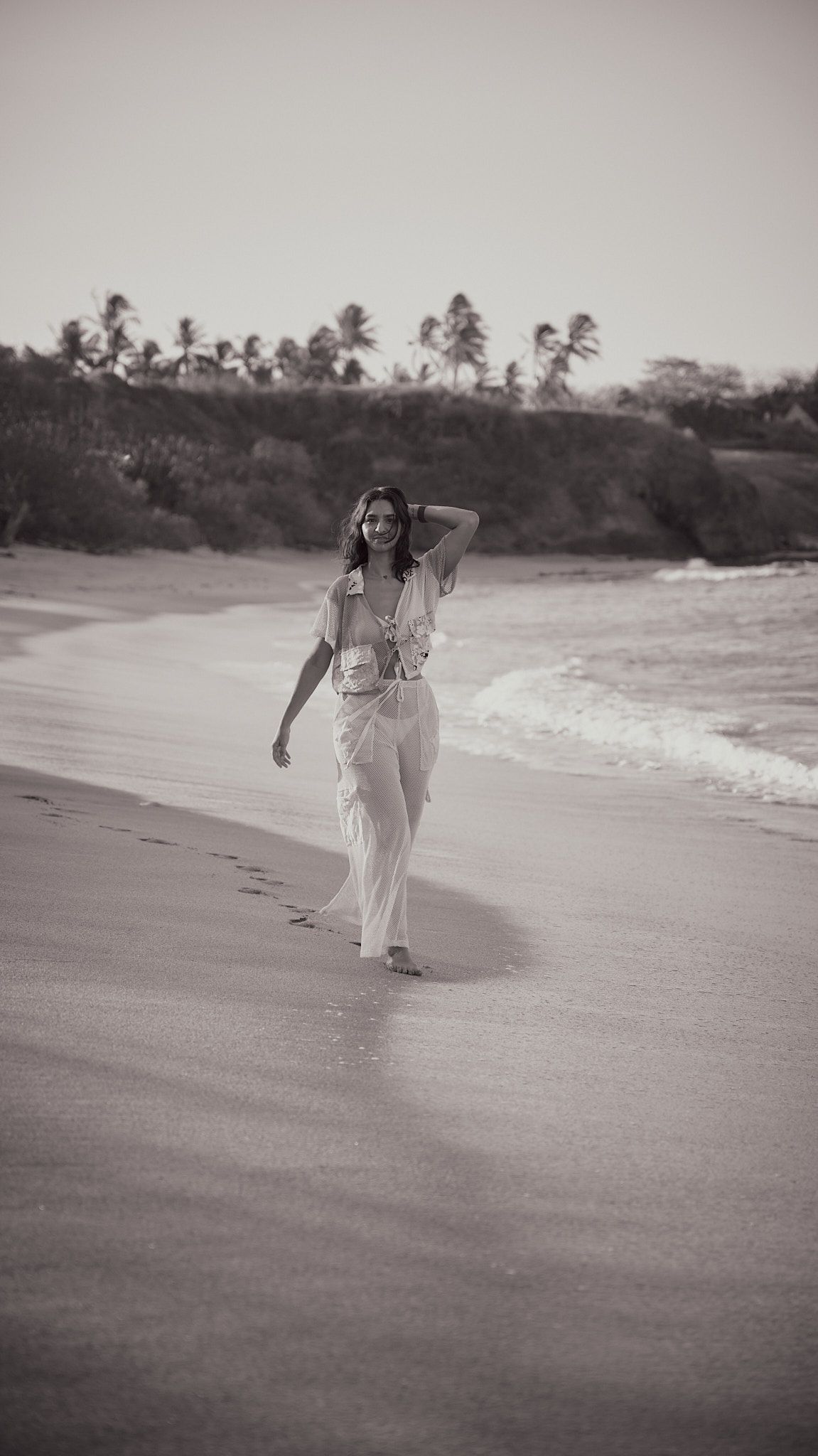 A person walking alone on a tropical beach in white clothing while palm trees line the horizon in black and white.