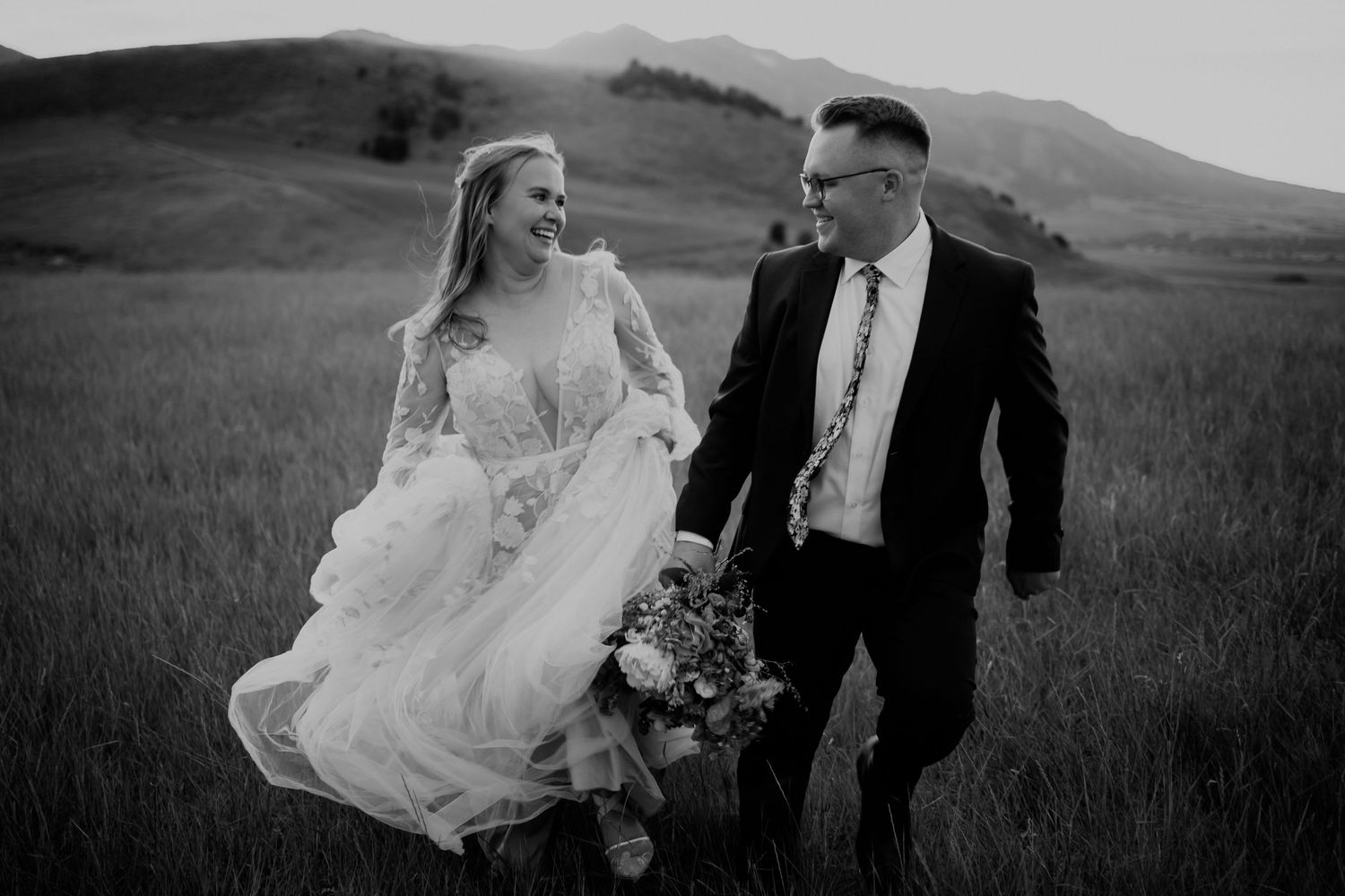 A black and white wedding portrait of a couple running through a scenic mountain meadow.