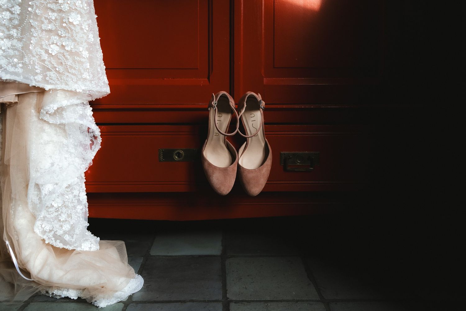 Pink bridal shoes hanging on burgundy cabinet next to white lace wedding dress in moody lighting.