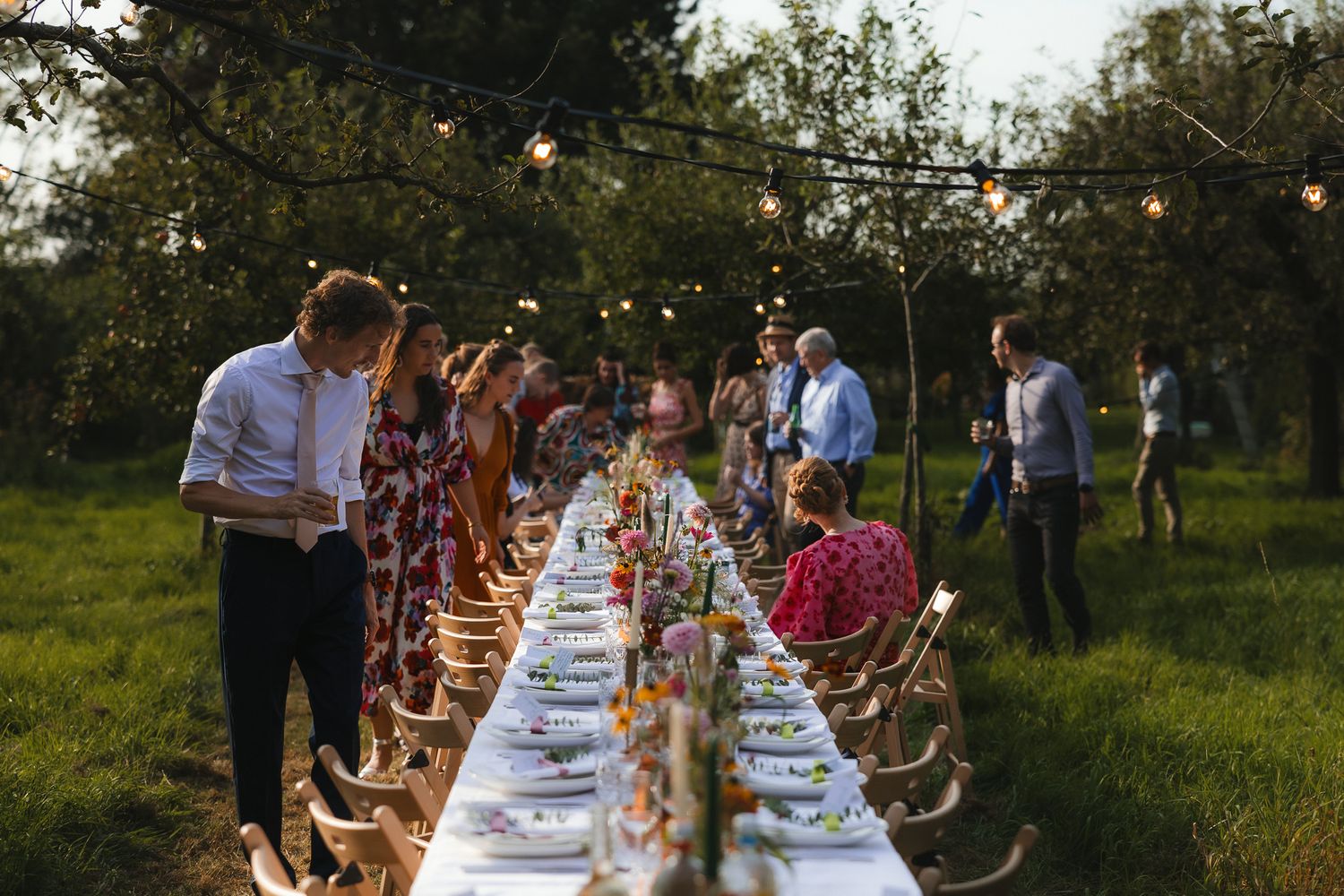 Long banquet table set for outdoor dinner party in garden with string lights and guests mingling at dusk.