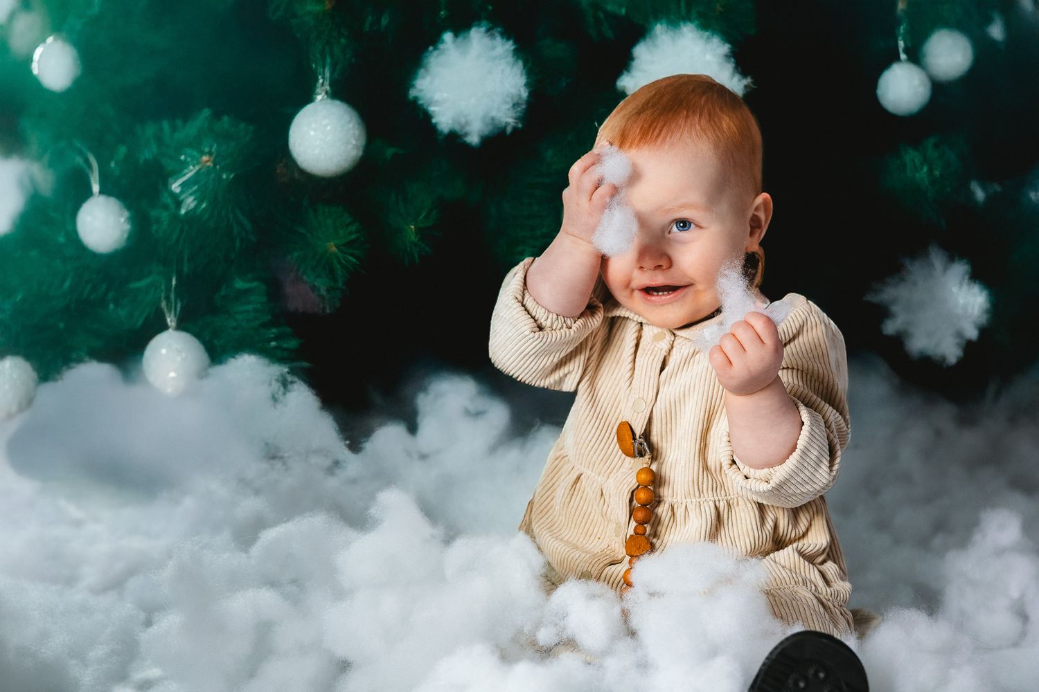 Baby wearing beige outfit sits in cloud-like setting with white ornaments and pom poms against dark green background.
