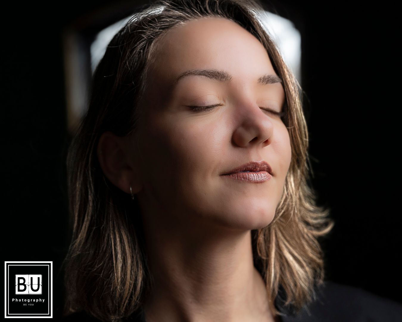 A woman with closed eyes and natural makeup poses serenely against a dark background.