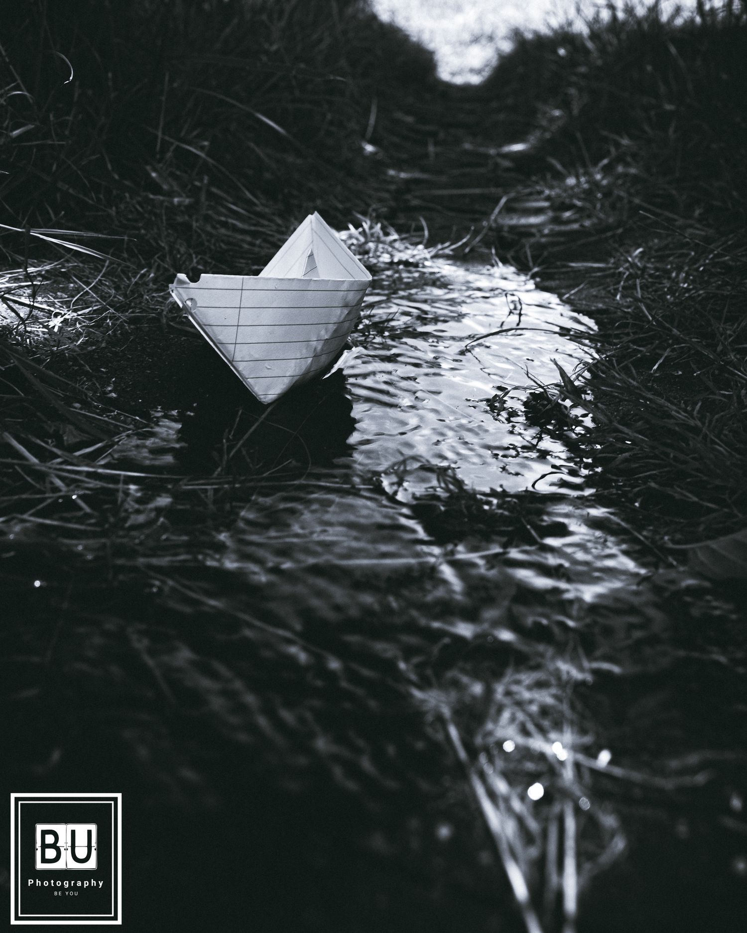 A black and white photograph of a paper boat floating on dark rippling water among tall grass.