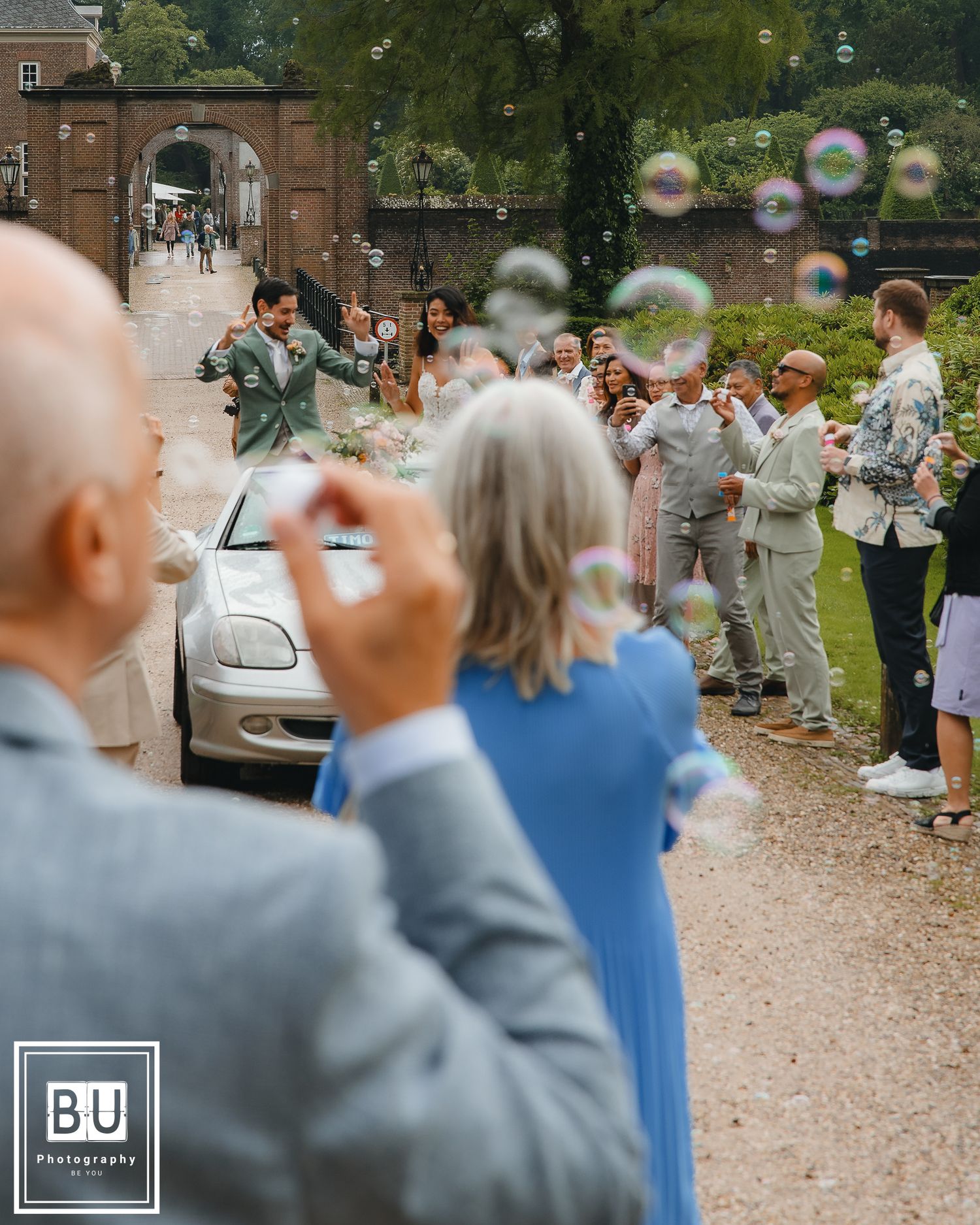 Wedding party stands along a path with bubbles floating in the air as guests blow them near a stone archway entrance.