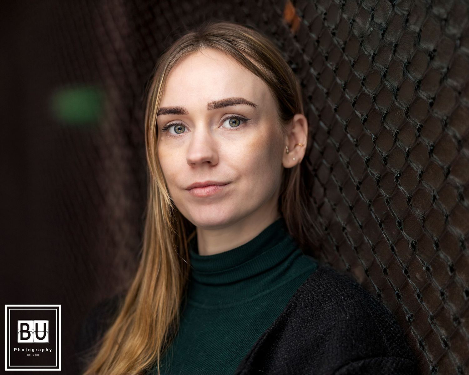 Professional portrait of a person with blonde hair wearing a dark green turtleneck and black sweater against a textured wall.