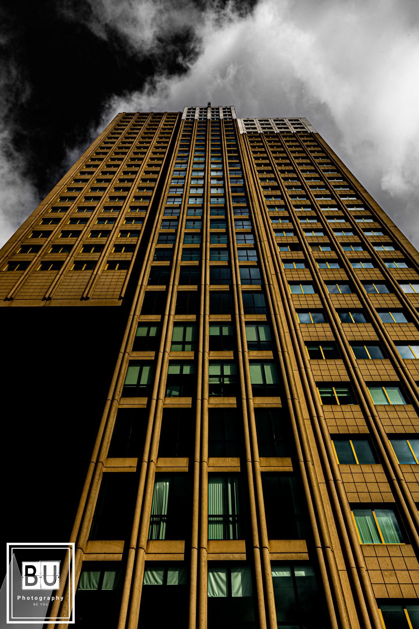 Dramatic upward view of a golden high-rise office building against dark cloudy skies with a BU watermark in corner.