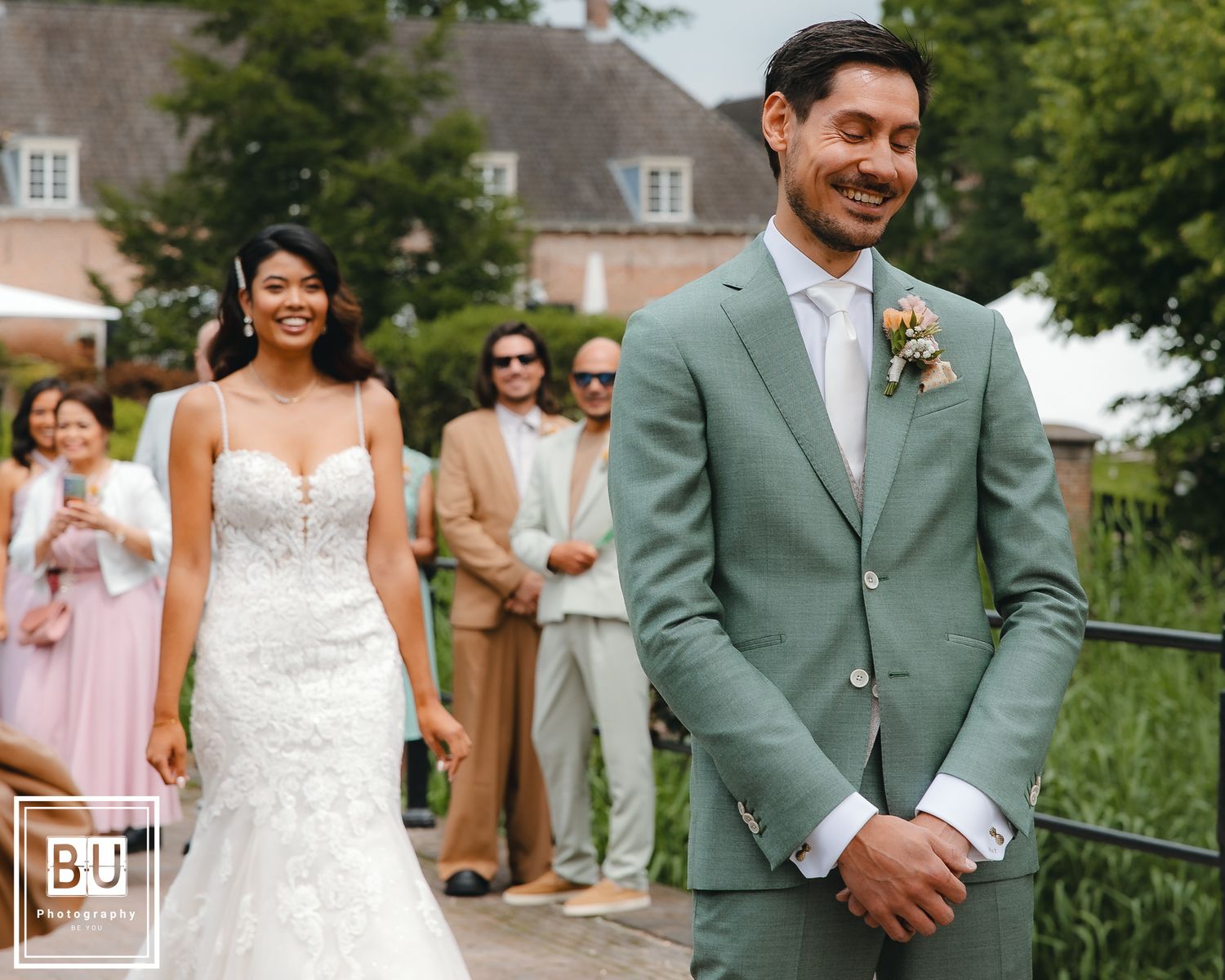 Wedding couple in elegant attire pose on brick pathway with wedding party in background.