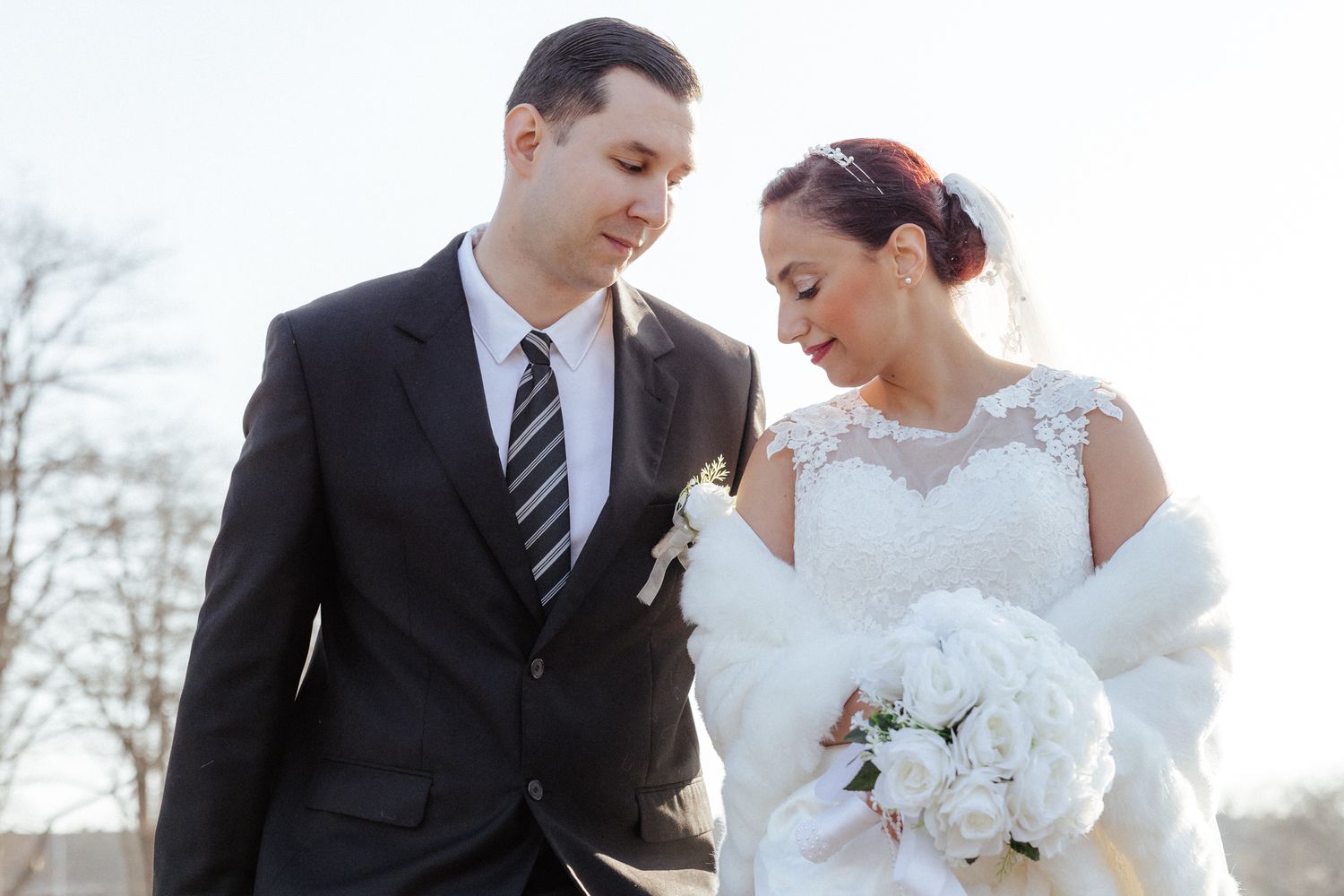 A bride and groom share an intimate moment outdoors while holding a white floral bouquet.
