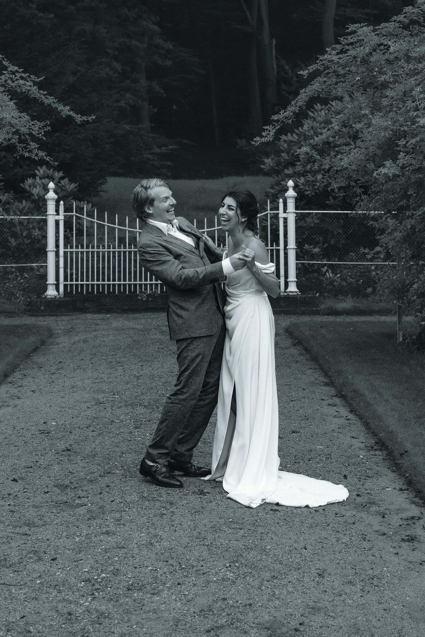 A black and white wedding photo of a couple dancing joyfully in front of an ornate metal gate in a garden setting.