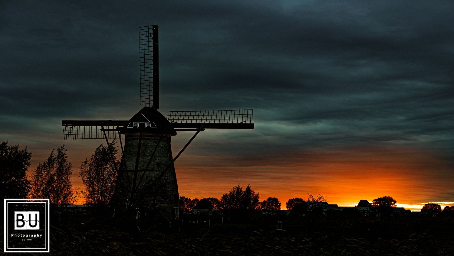 Traditional Dutch windmill silhouetted against dramatic sunset sky with dark storm clouds and glowing orange horizon.