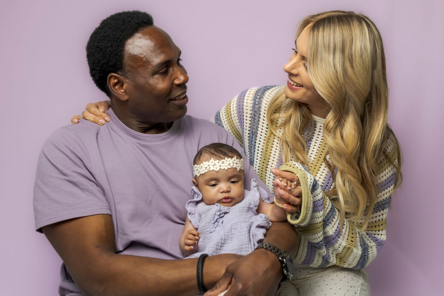 A family poses together against a light background, with a baby wearing a headband in the middle.