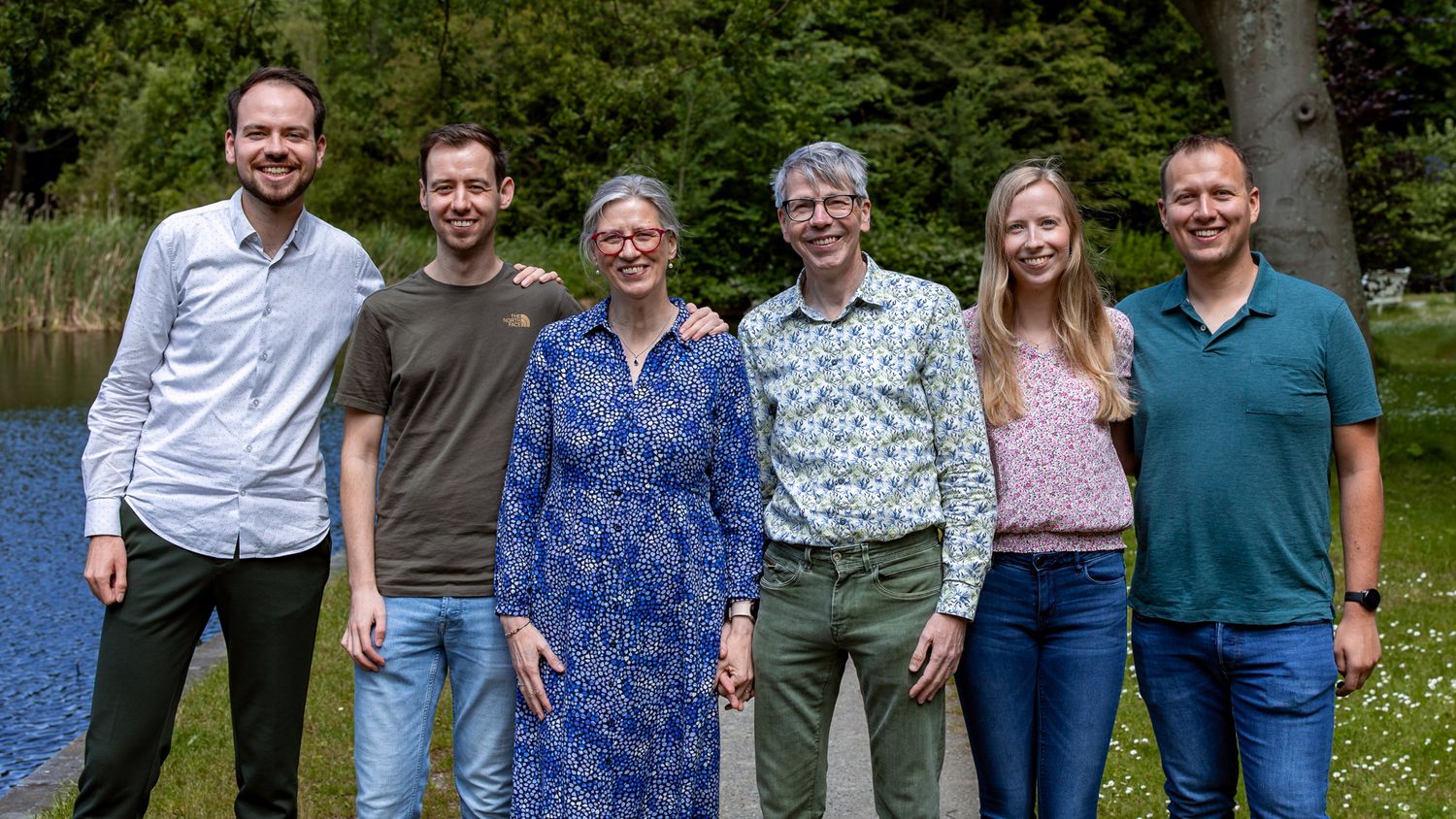 A group of six people standing together and smiling outdoors by a lake on a sunny day.