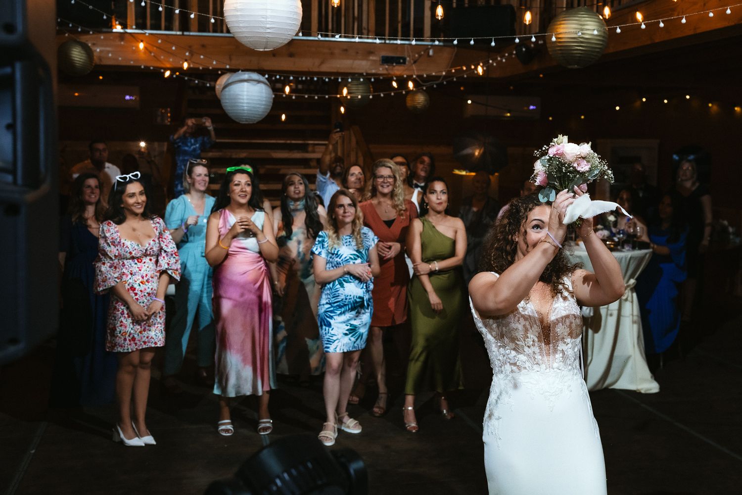 Wedding guests watch as bride tosses bouquet during reception in rustic venue with string lights overhead.