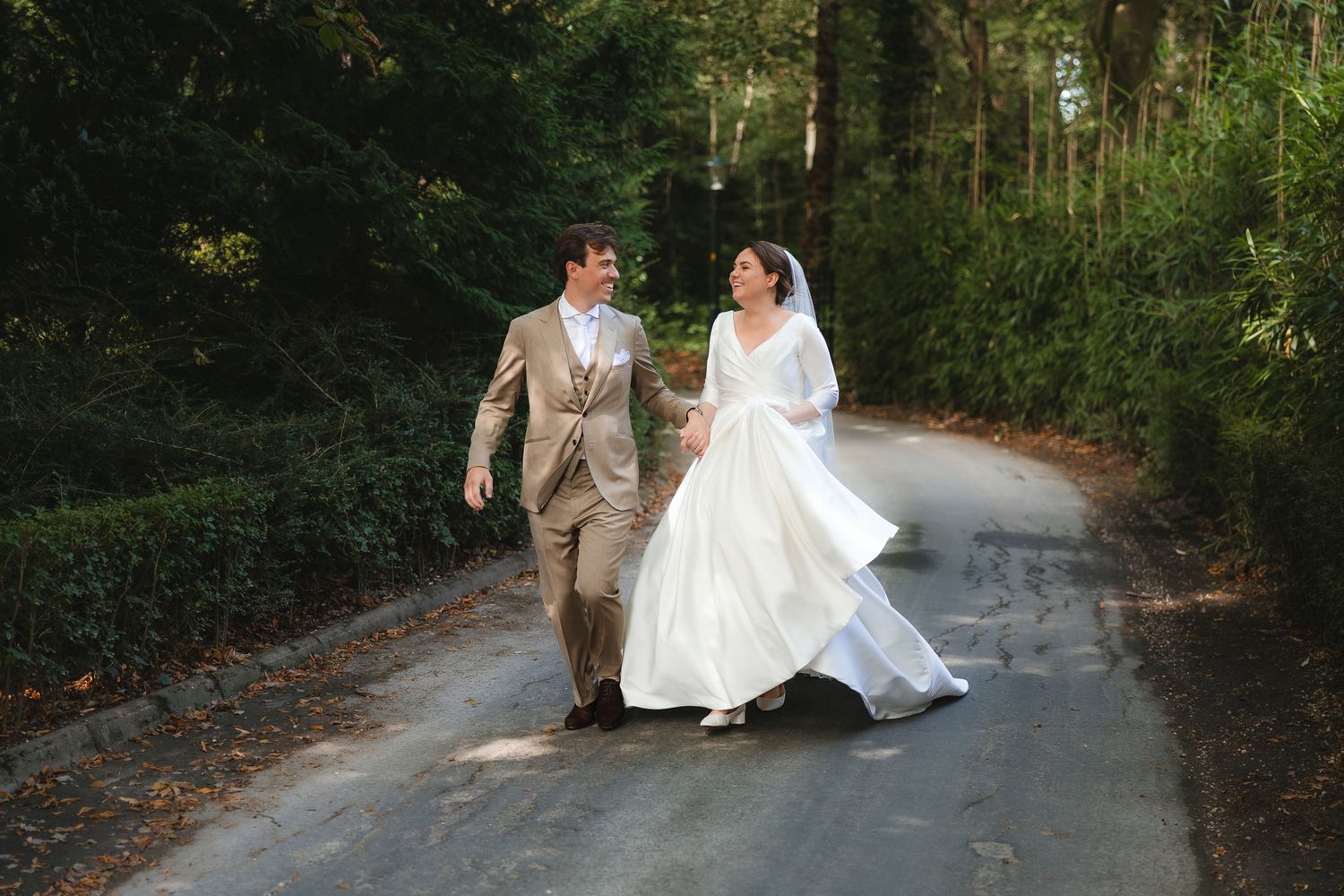 A happy couple in wedding attire walks along a scenic forest road as the bride's dress flows in the breeze.