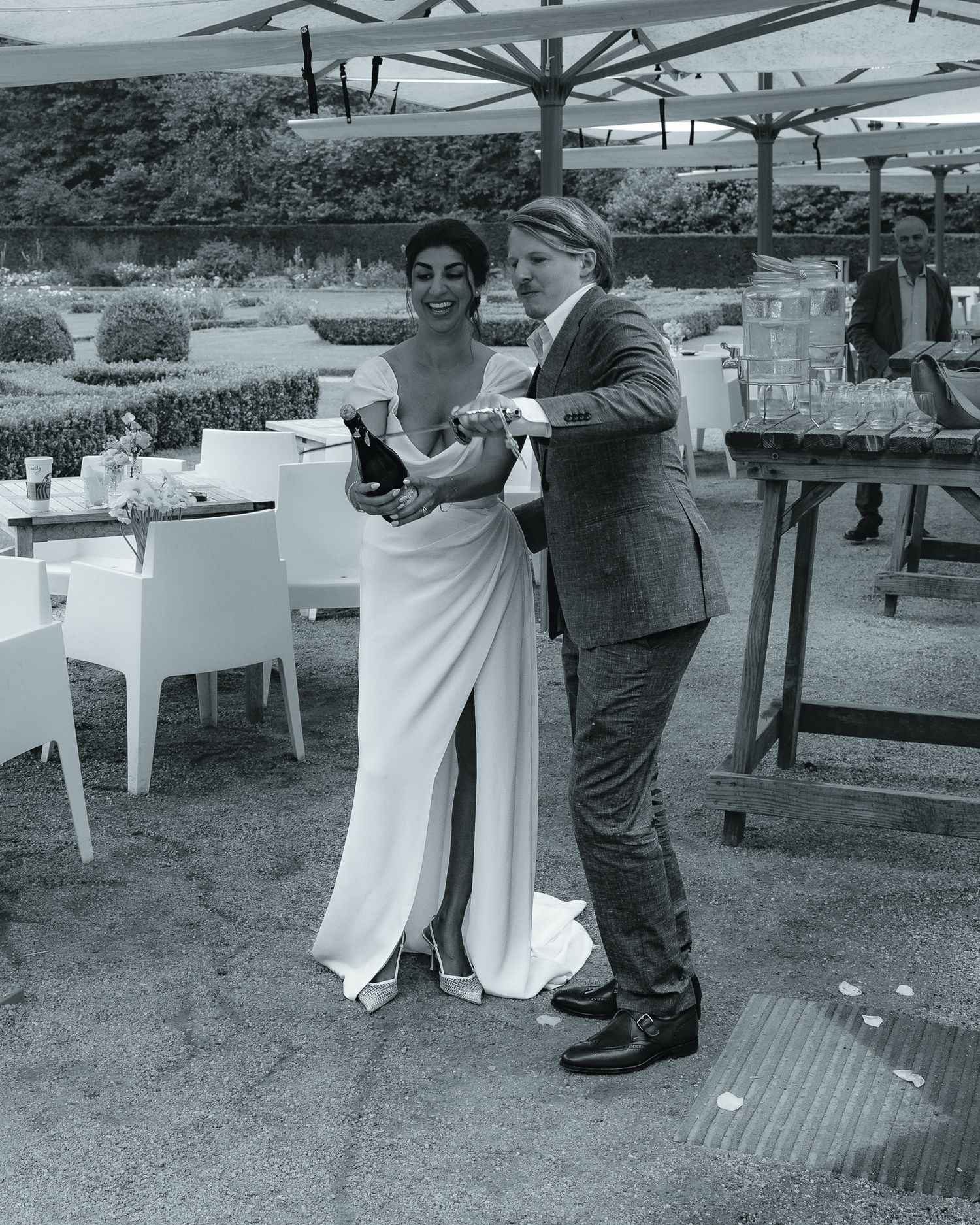 A black and white candid image of an outdoor wedding reception with guests interacting under patio umbrellas.