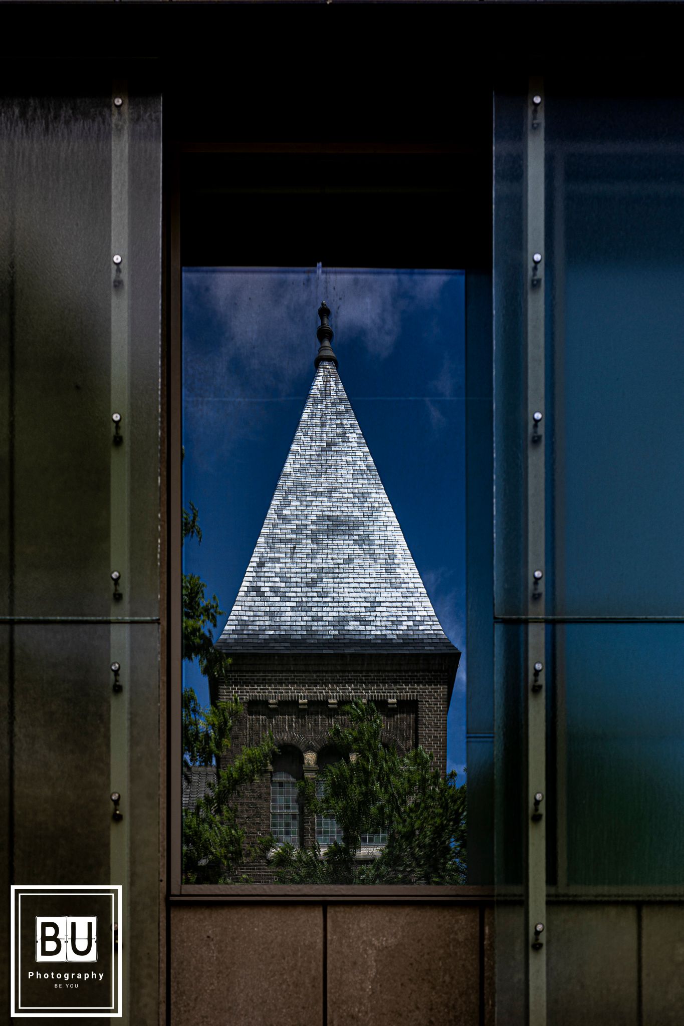 A stone church spire with a triangular white top is framed between modern building walls against a blue sky.