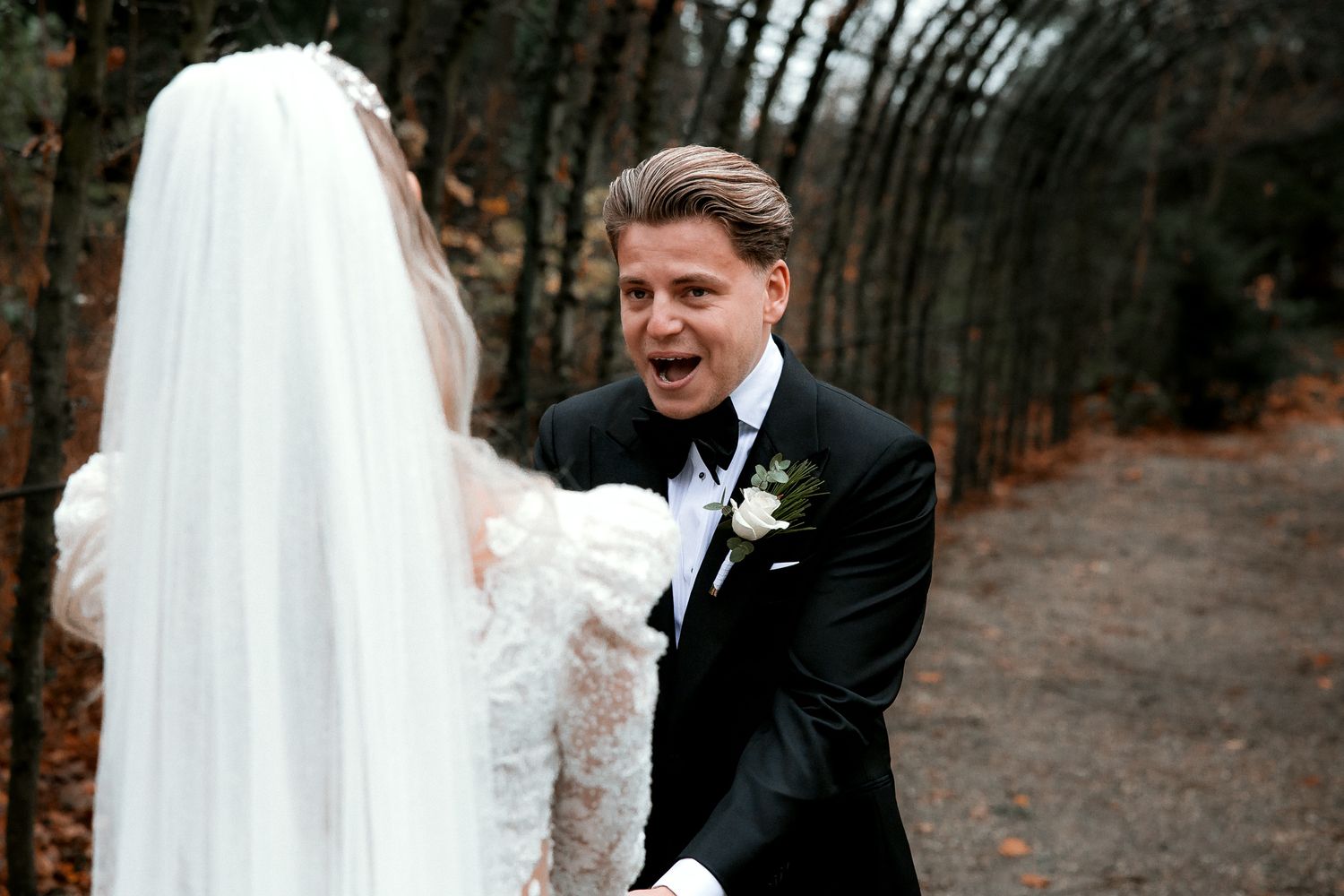 A joyful wedding moment between a bride in white lace and groom in black suit along a wooded garden path.